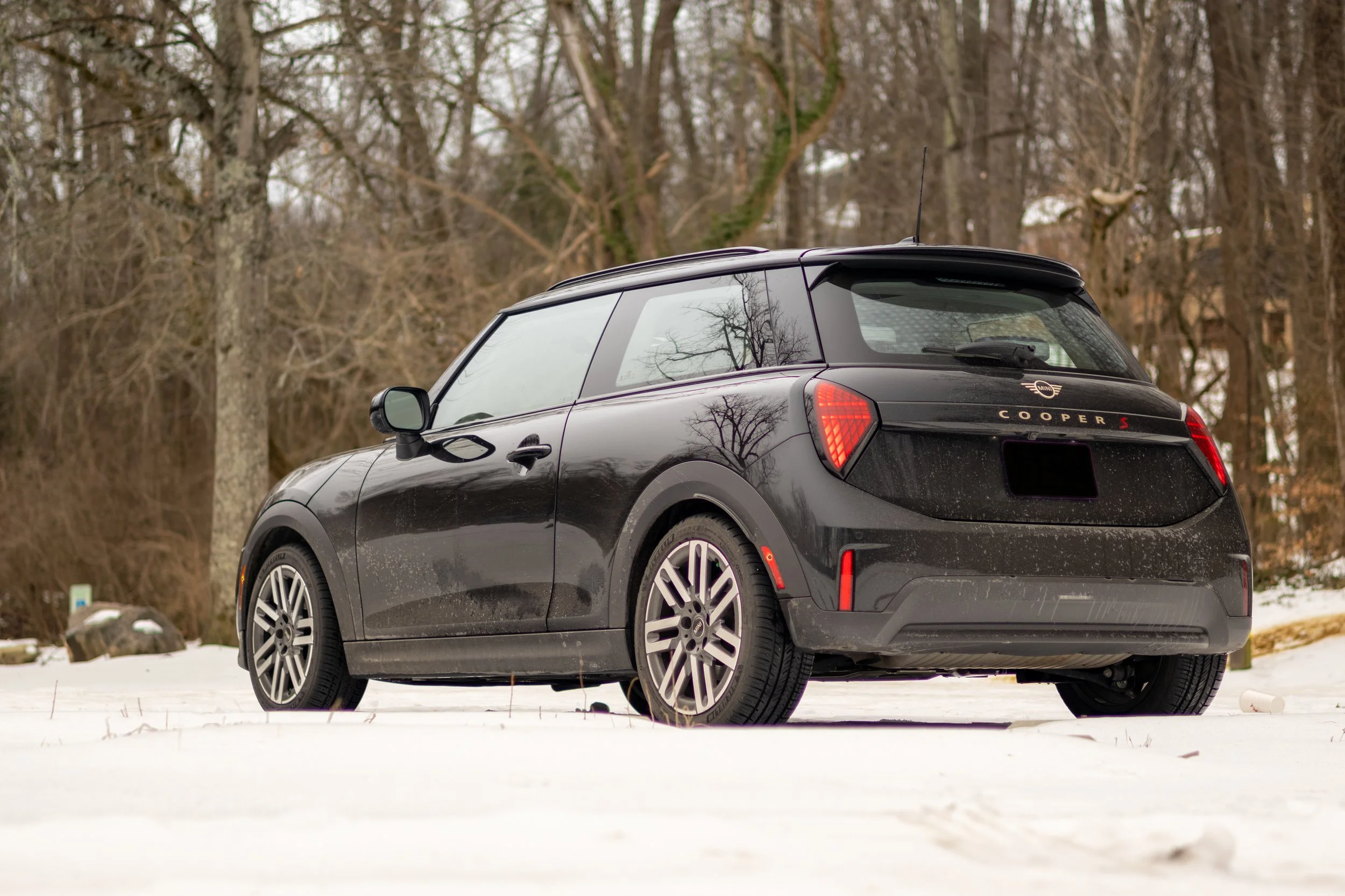 A black Mini Cooper S parked on snow-covered ground with leafless trees in the background. Car meet. Cars and coffee. Rollers. Stills. Detail shots. Brand awareness. MINI Cooper S
