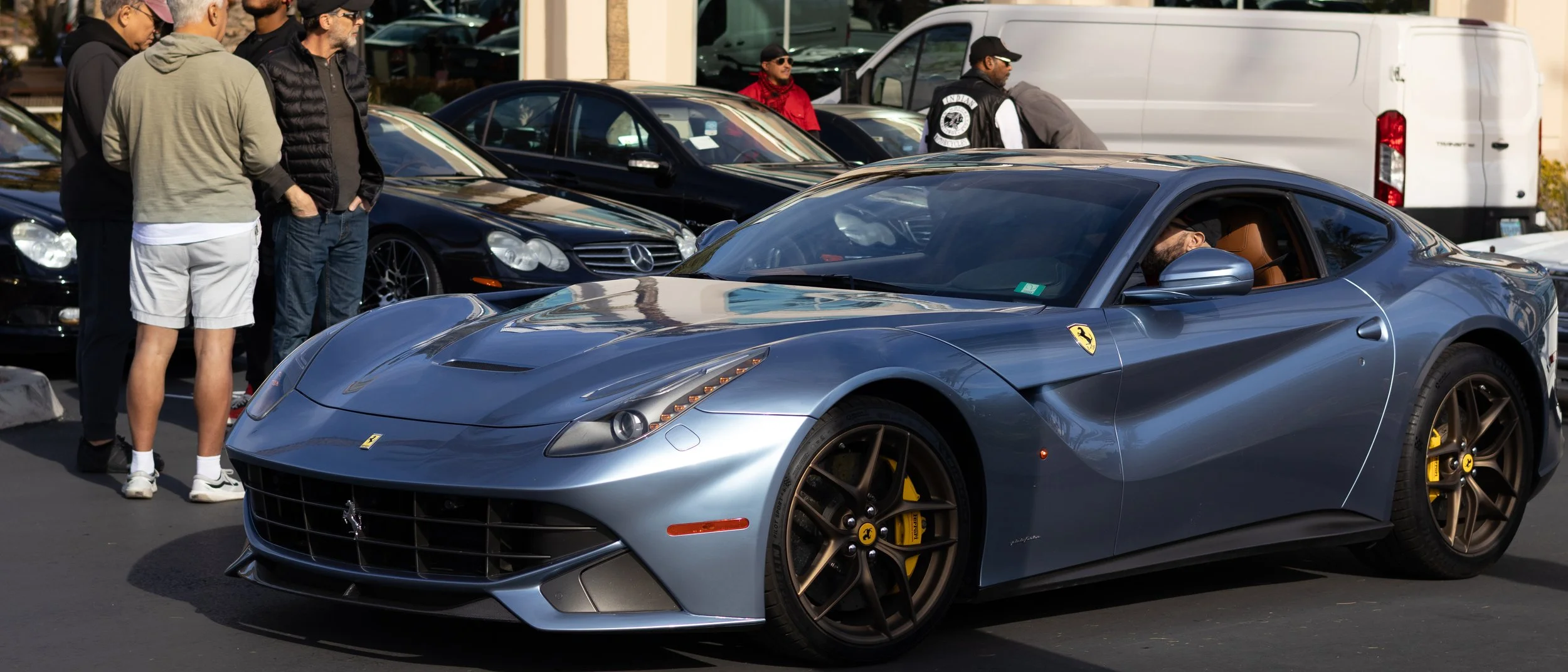 A silver Ferrari sports car parked at an outdoor car show with several people gathered around, including a man inside the car, and in the background, there are other luxury cars and a white van. F12.