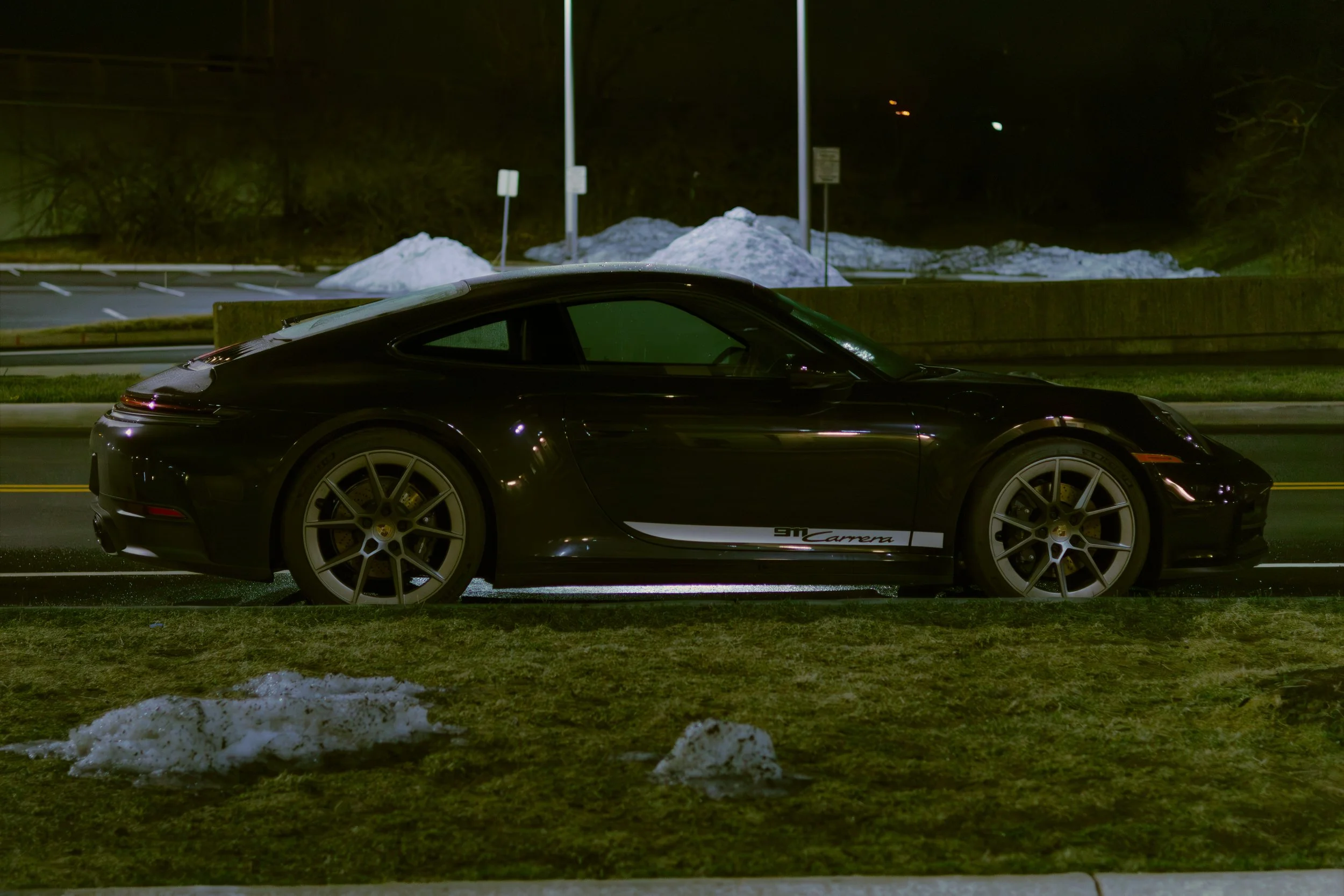 A black Porsche 911 Carrera parked on the side of a street at night, with snow piles and a parking lot in the background. Car meet. Cars and coffee. Rollers. Stills. Detail shots. Brand awareness. Porsche 911.
