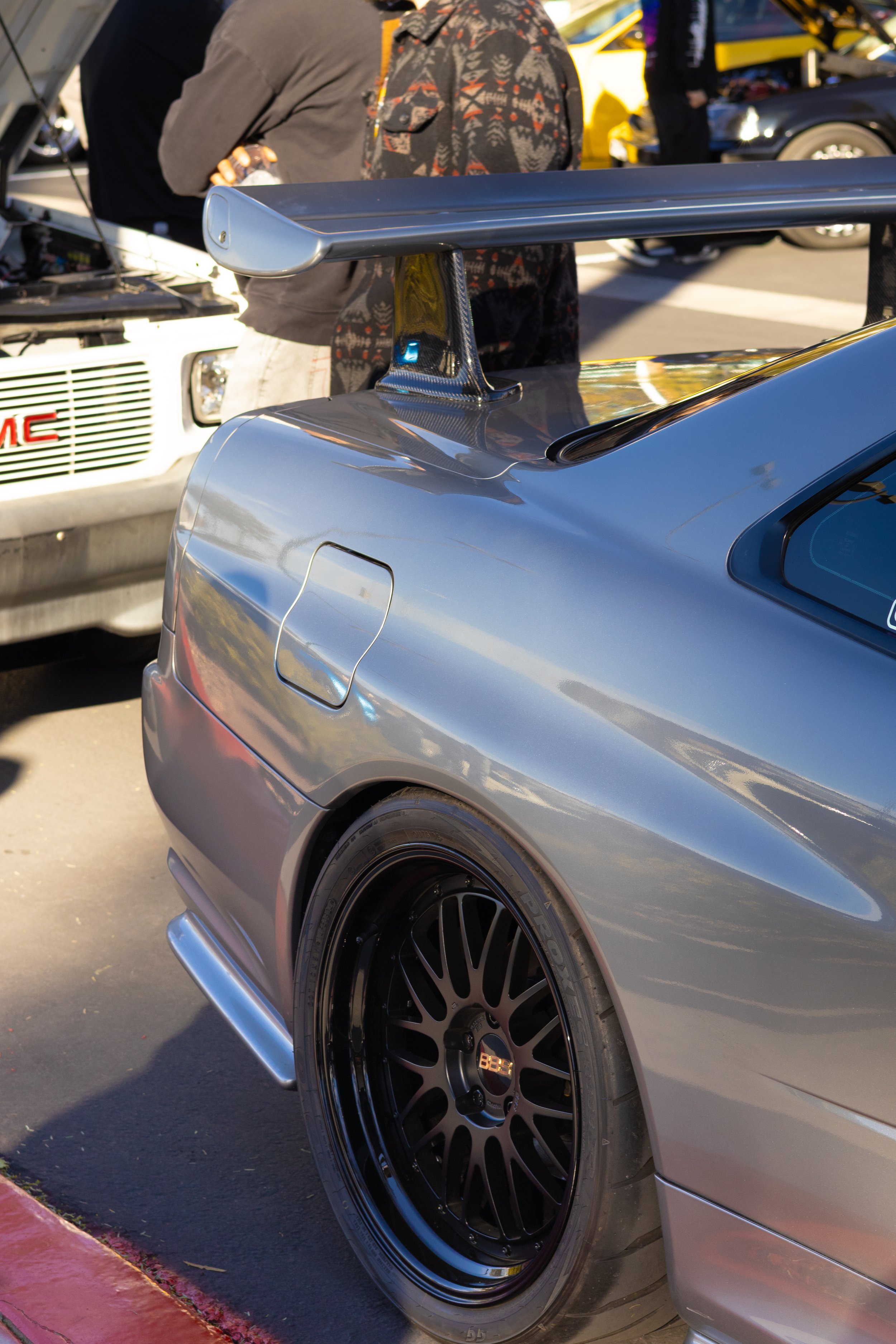 Close-up of the rear side of a silver sports car with a large rear wing, black wheels, and a decal on the window, parked at an outdoor event with people and classic cars in the background. Nissan R34
