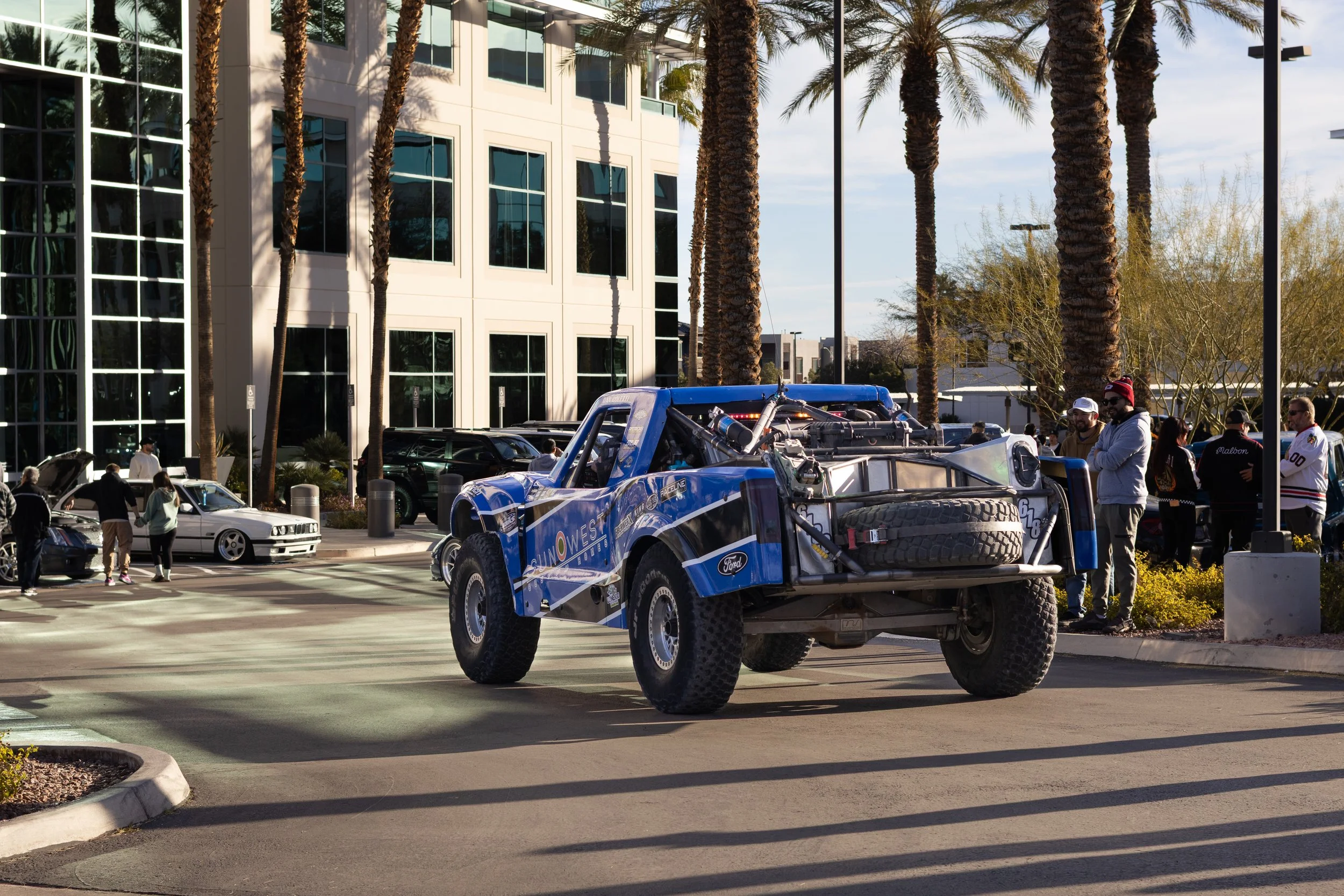 A blue off-road racing truck with large tires, parked in a parking lot with palm trees and a modern building in the background. Several people are observing the truck and cars nearby. Trophy Truck. BAJA 1000.