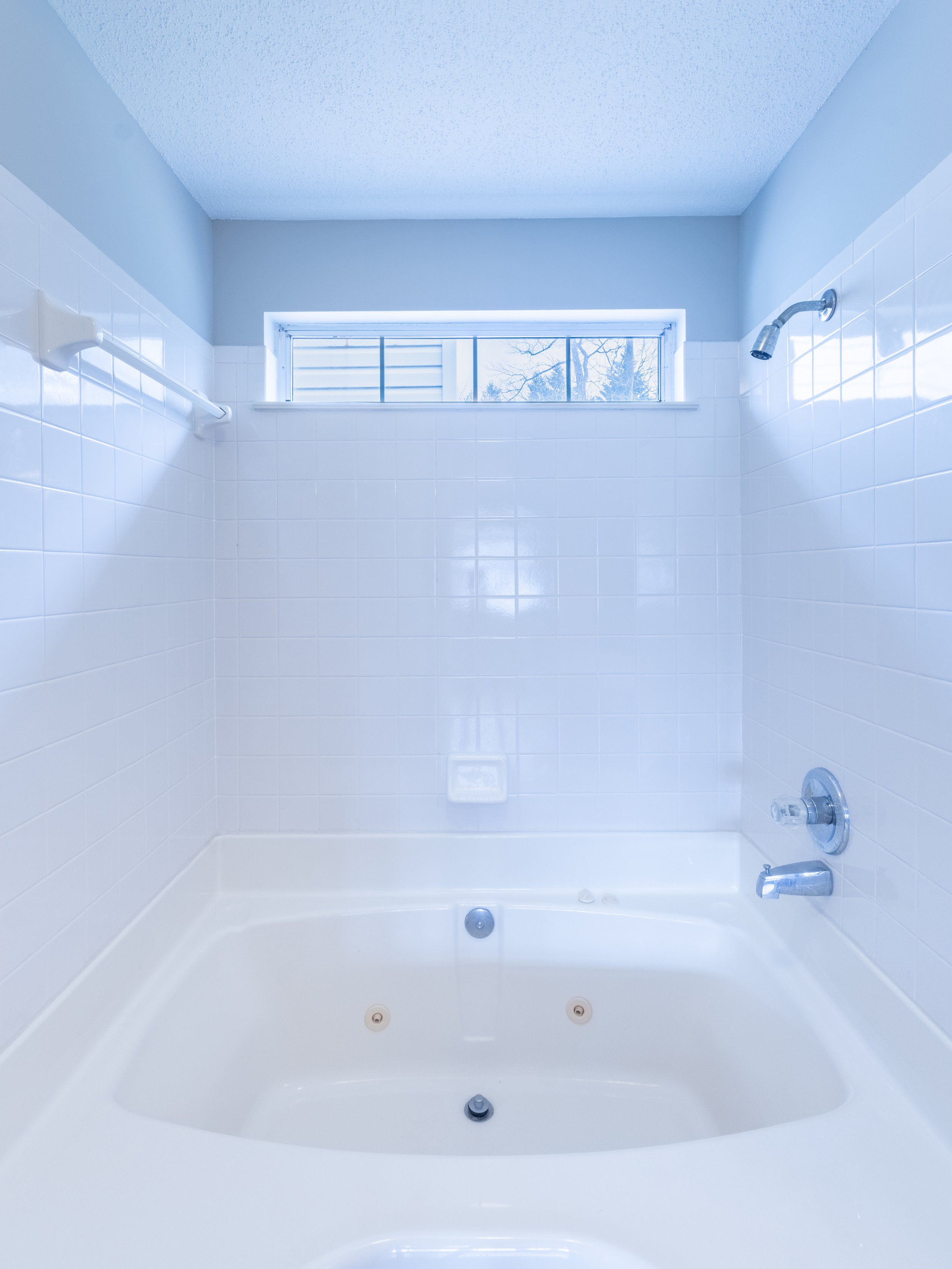 Empty bathroom with bathtub, white tiled walls, a small window high on the wall, and a showerhead.