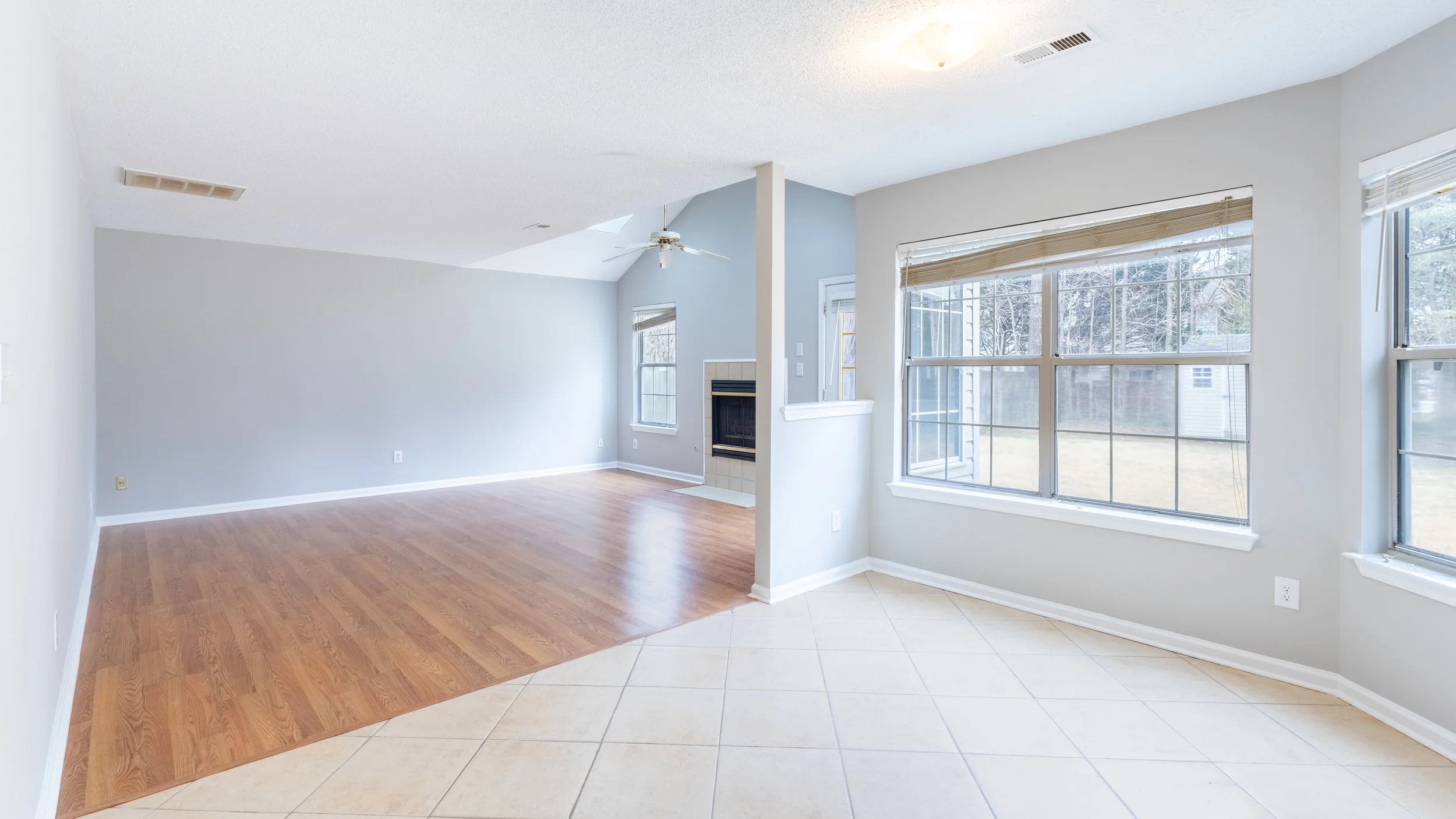 Empty living room with large windows, wood and tile flooring, fireplace, and ceiling fan.