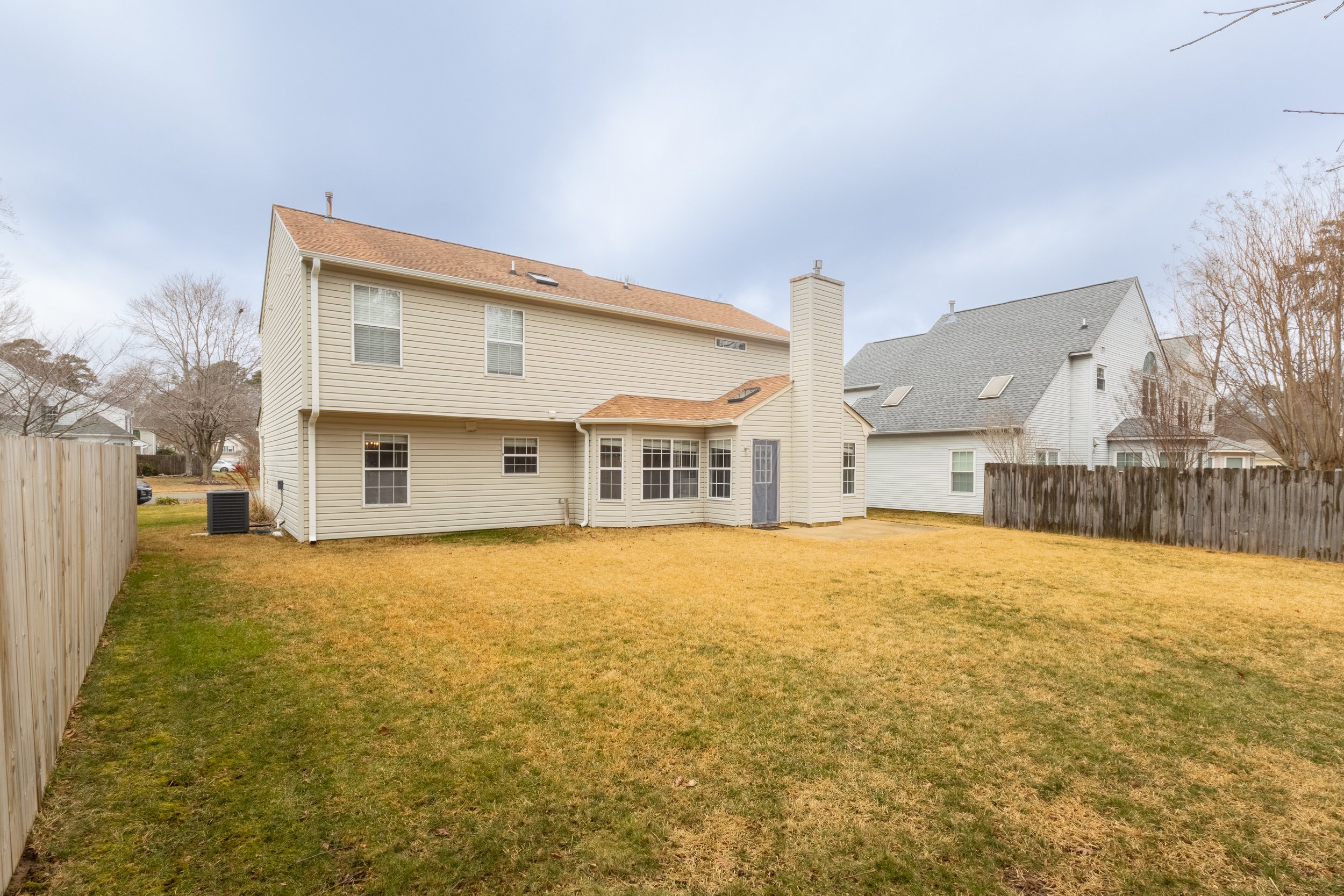 Backyard of a suburban house with tan siding, multiple windows, a chimney, and a fenced yard with grass, during overcast weather.