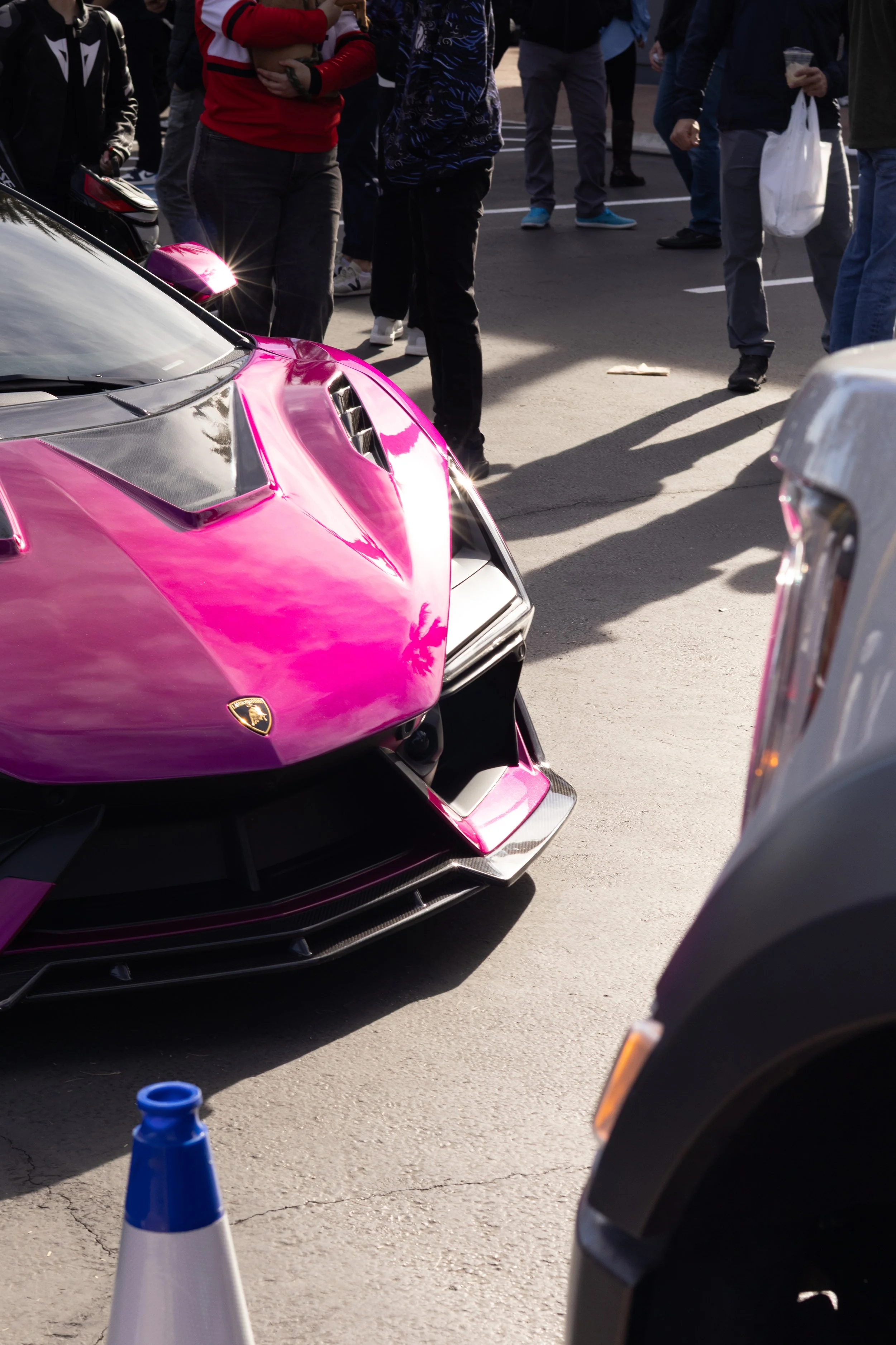 Close-up of a shiny pink and black Lamborghini parked on a city street, with a crowd of people standing and walking nearby. Car meet. Cars and coffee. Rollers. Stills. Detail shots. Brand awareness. 