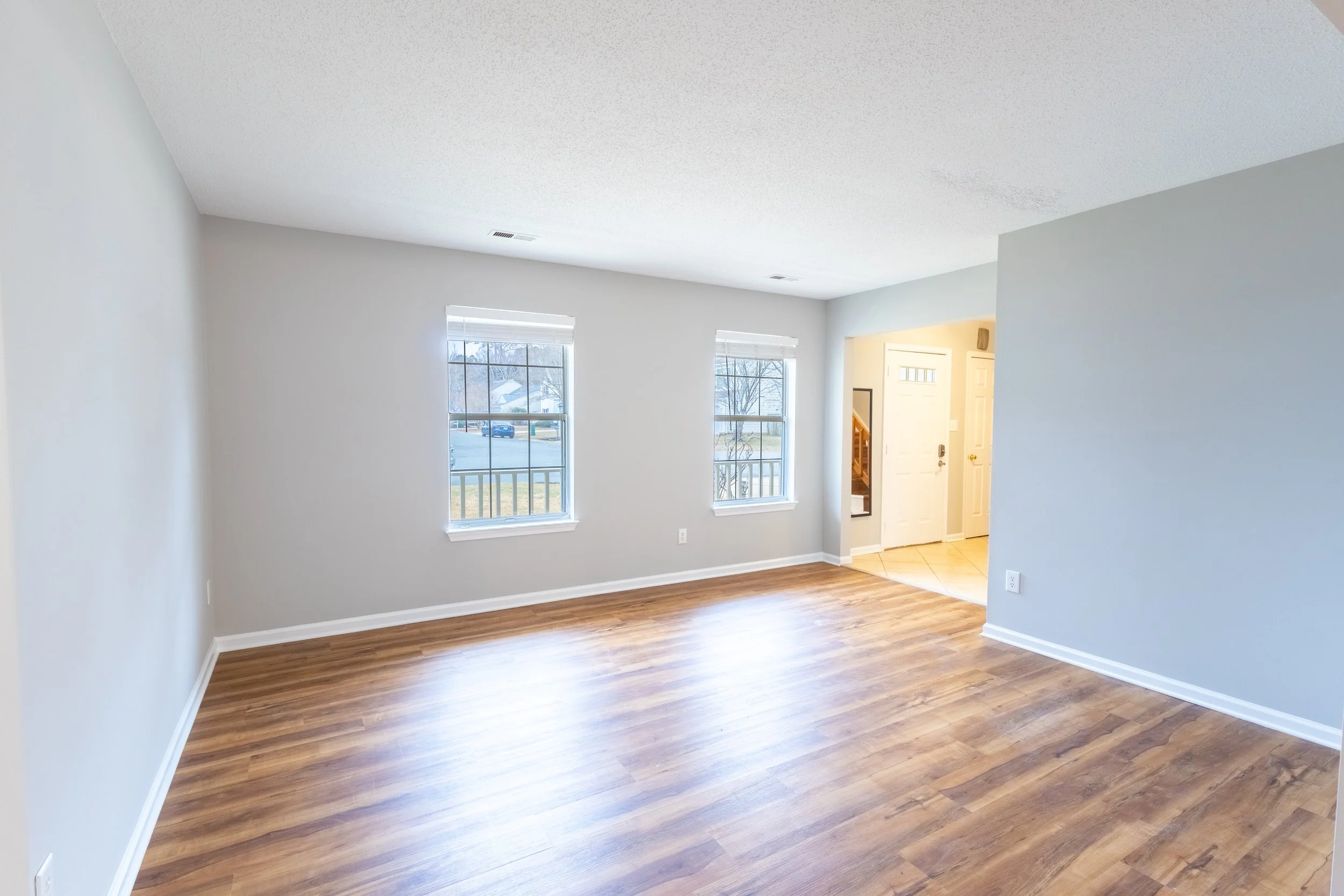 Empty living room with hardwood floors, light gray walls, two windows, and an open entryway to the front door area.