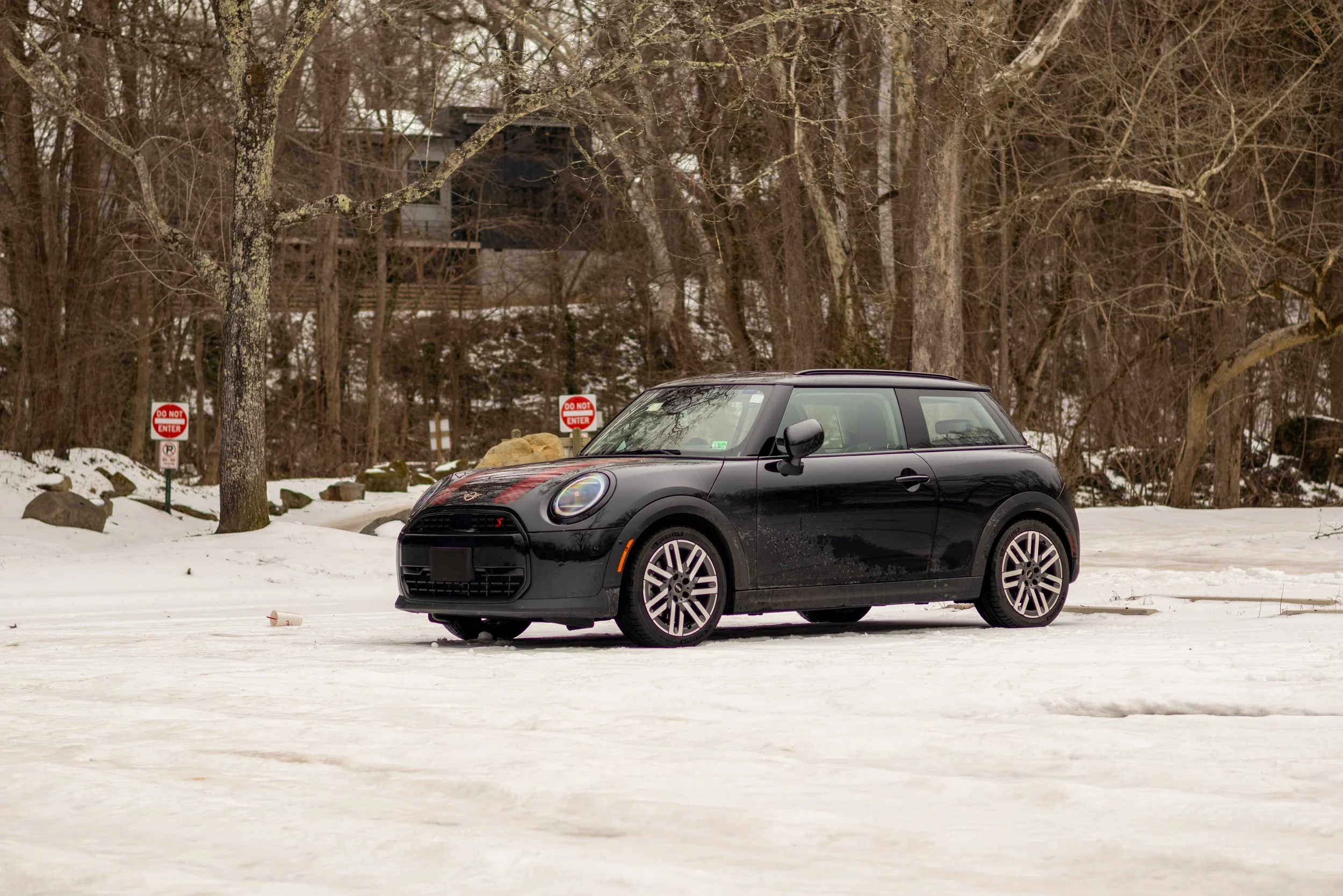 A black car parked on a snow-covered road in a wooded area with several 'Do Not Enter' signs in the background. Car meet. Cars and coffee. Rollers. Stills. Detail shots. Brand awareness. MINI Cooper S.