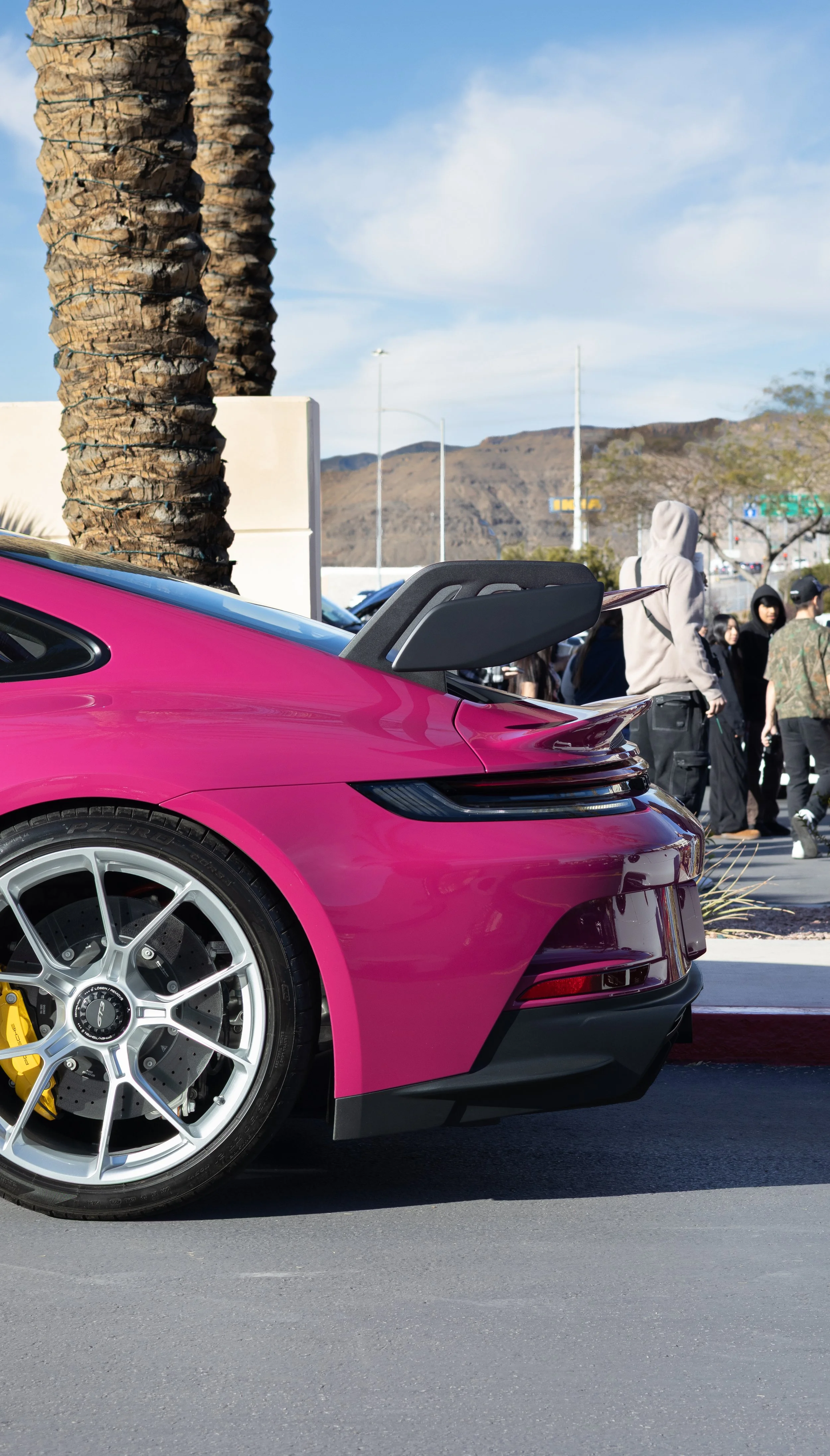 Close-up of the rear side of a pink sports car with a black spoiler, parked on a city street with palm trees, people walking, and mountain background. Car meet. Cars and coffee. Rollers. Stills. Detail shots. Brand awareness. Porsche. GT3