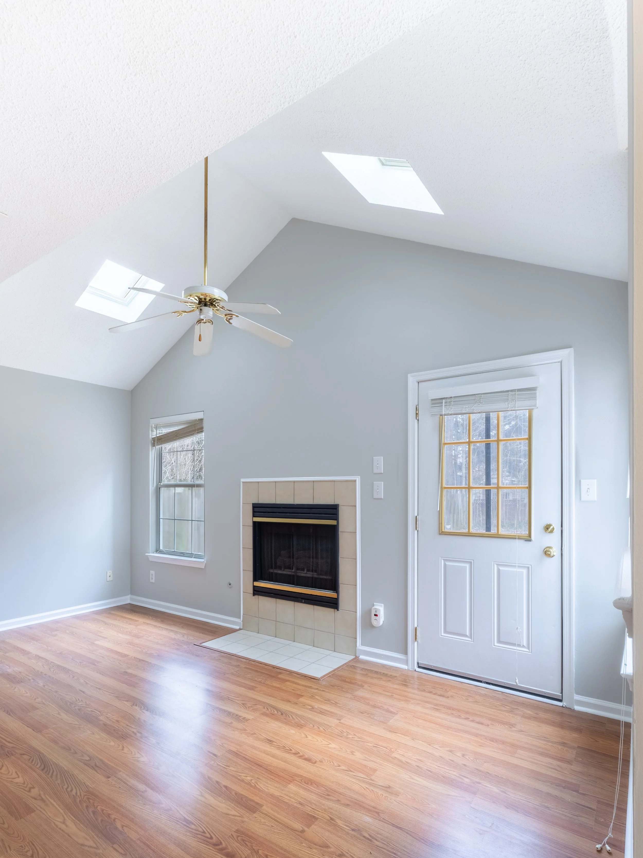 Empty room with light gray walls, hardwood floor, fireplace, ceiling fan, and skylights.