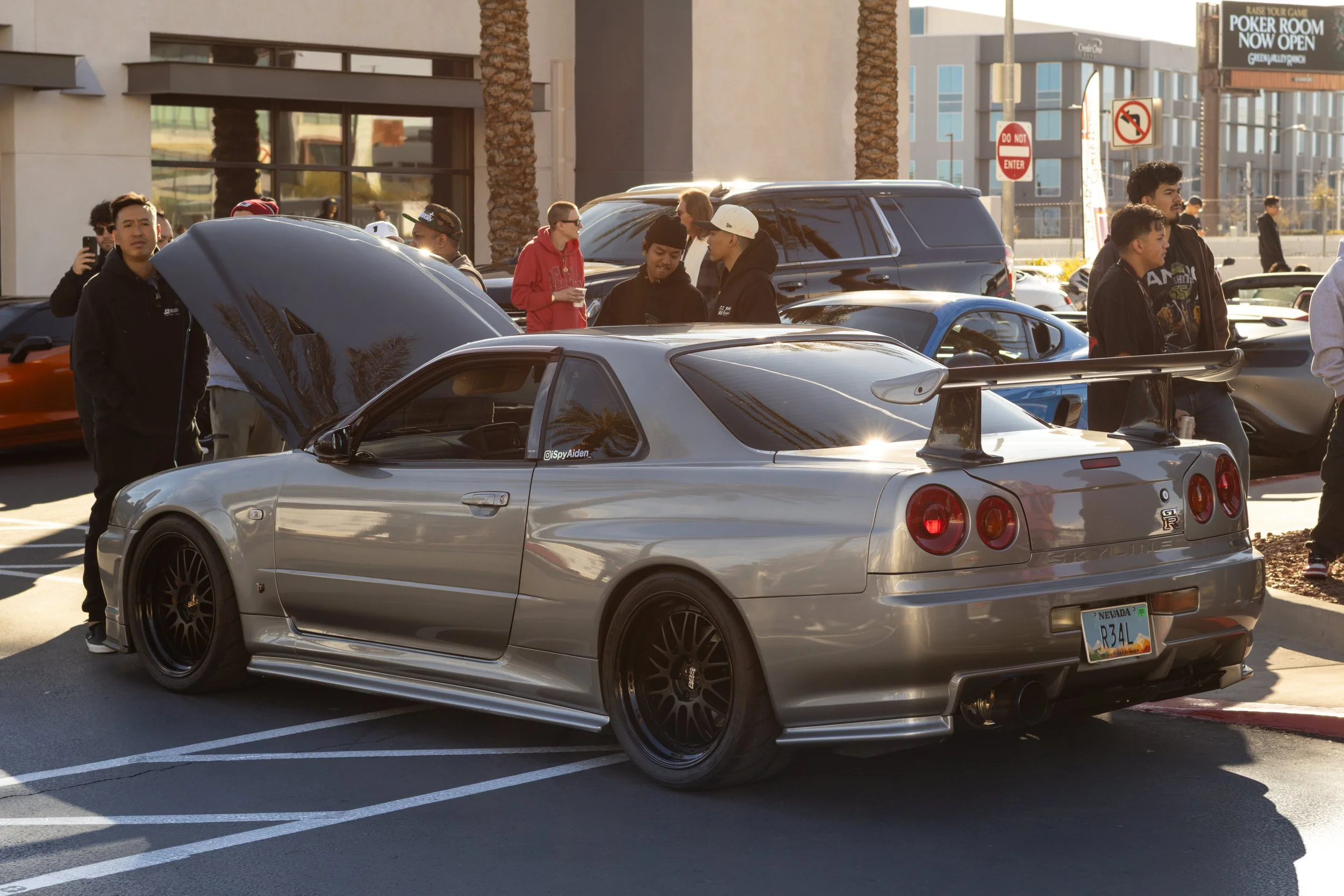 A silver R34 GTR sports car with a large rear wing and black wheels is parked in a lot with its hood open, surrounded by several people, some taking pictures or talking, in front of an urban building with palm trees and signs. 