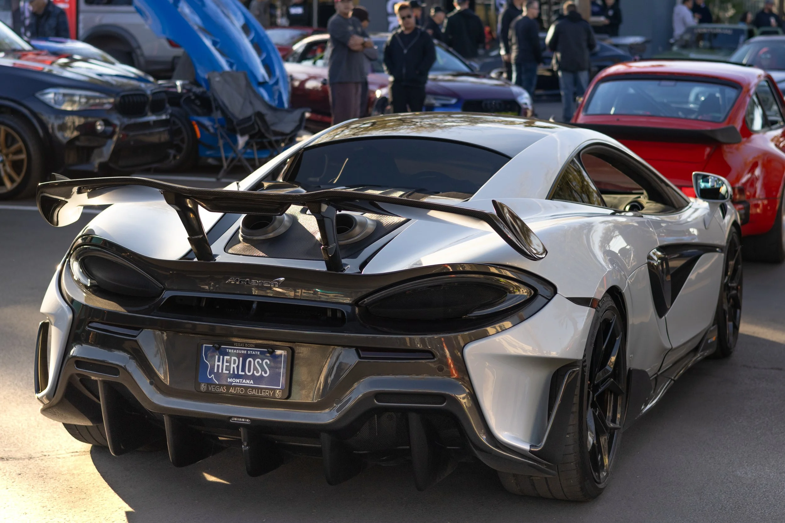 A white sports car with black accents and a large rear wing parked at an outdoor car show, with people and other cars in the background. Car meet. Cars and coffee. Rollers. Stills. Detail shots. Brand awareness.  Mclaren. 600LT.