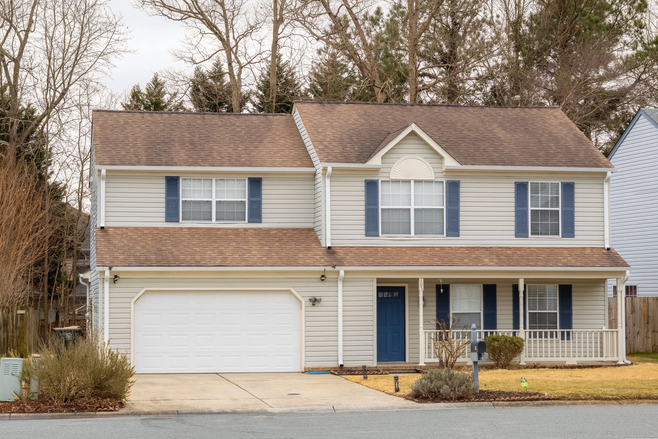 A two-story suburban house with beige siding, blue shutters, and a brown shingled roof. It has a front porch with white railing, a blue front door, and a white garage door. There are some bushes and shallow landscaping in front, with a mailbox near t