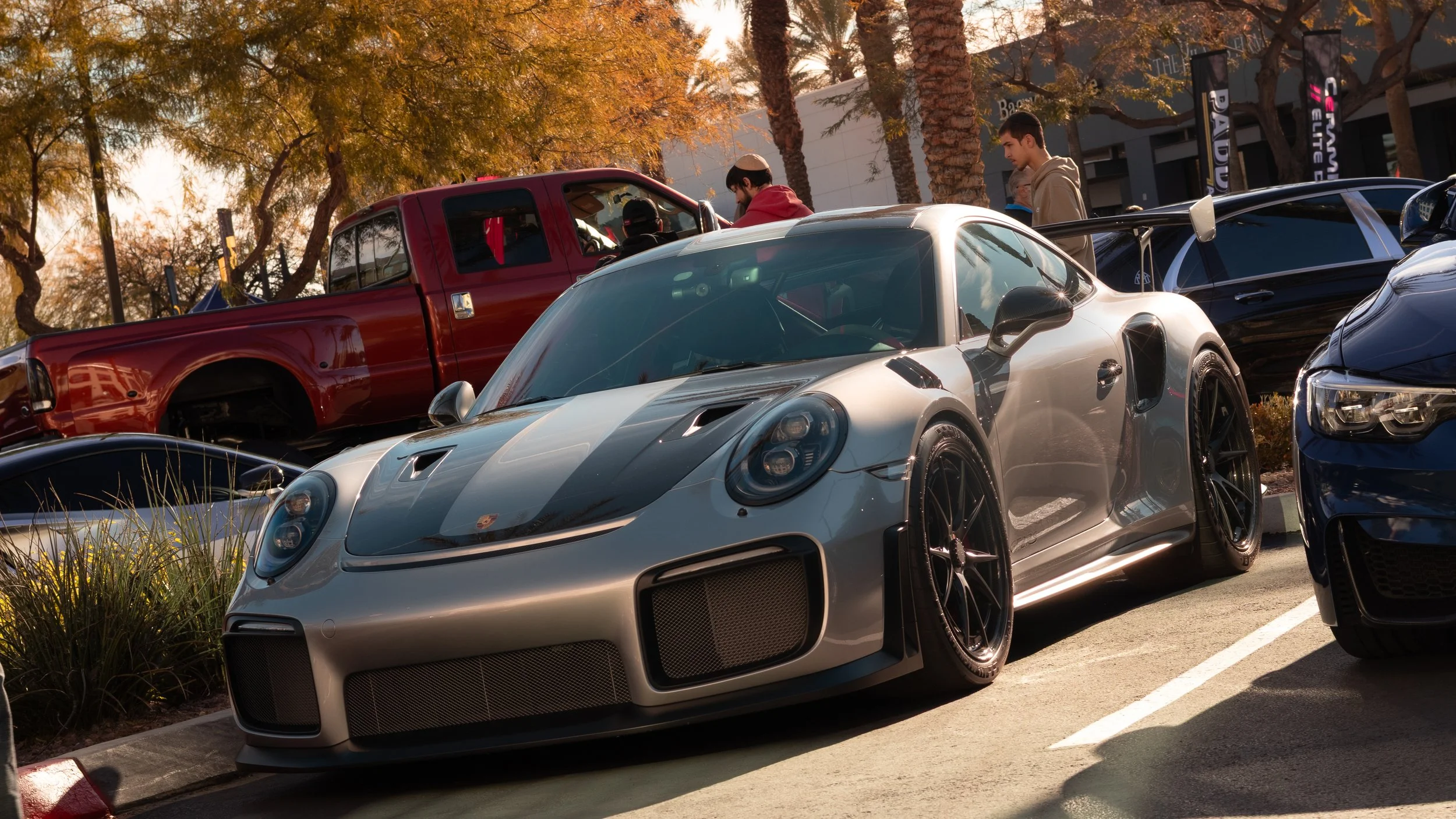 A silver Porsche sports car with a black racing stripe parked in a lot during what appears to be late afternoon or early evening, with a red pickup truck and several people in the background. GT2RS. 
