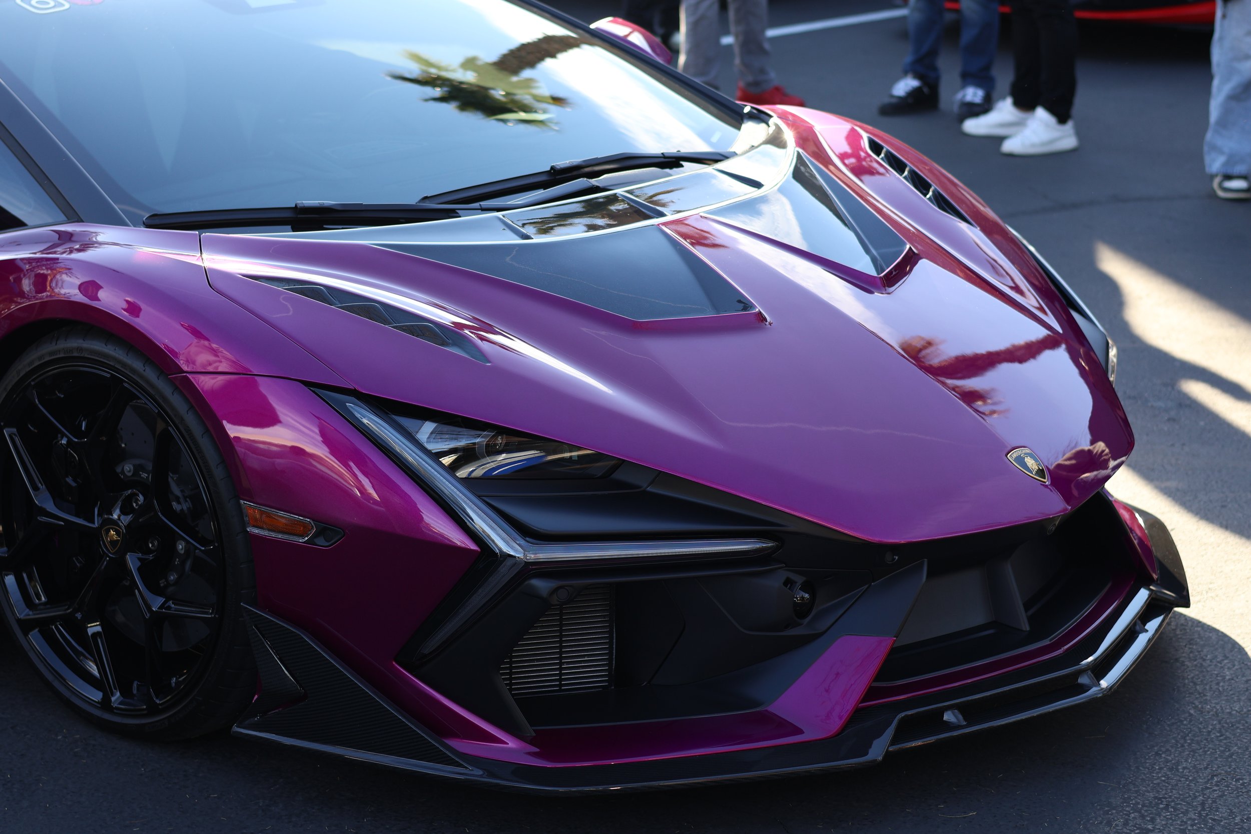 A close-up of the front part of a purple and black Lamborghini sports car parked outdoors, with people standing in the background. Car meet. Cars and coffee. Rollers. Stills. Detail shots. Brand awareness. 