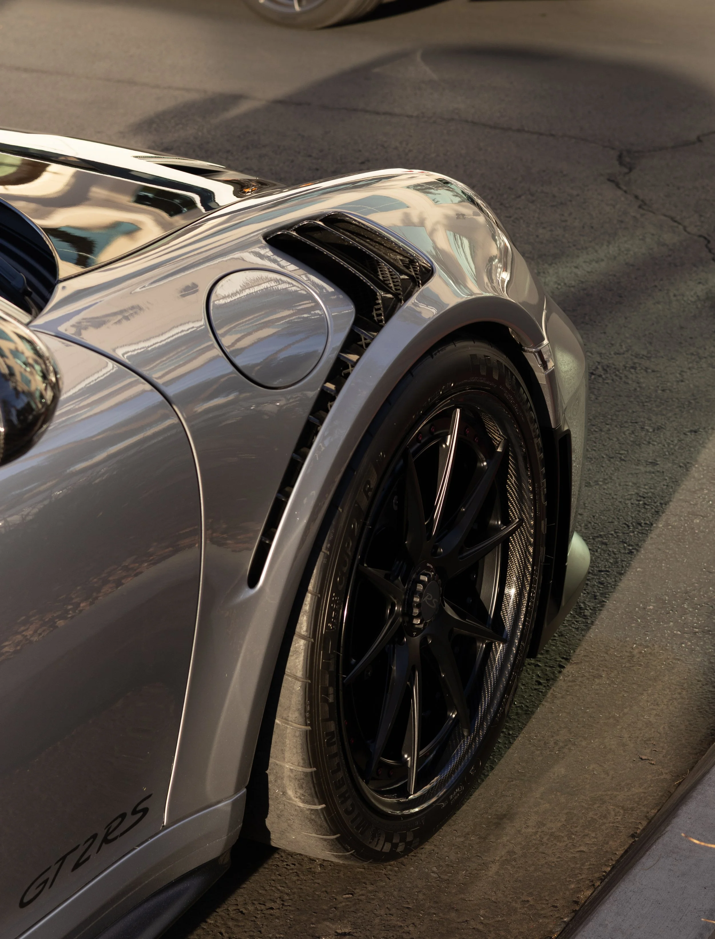 Close-up of a silver sports car's front right wheel, fender, and headlight, showing black alloy rims, tire, and aerodynamic details. Porsche GT2RS.