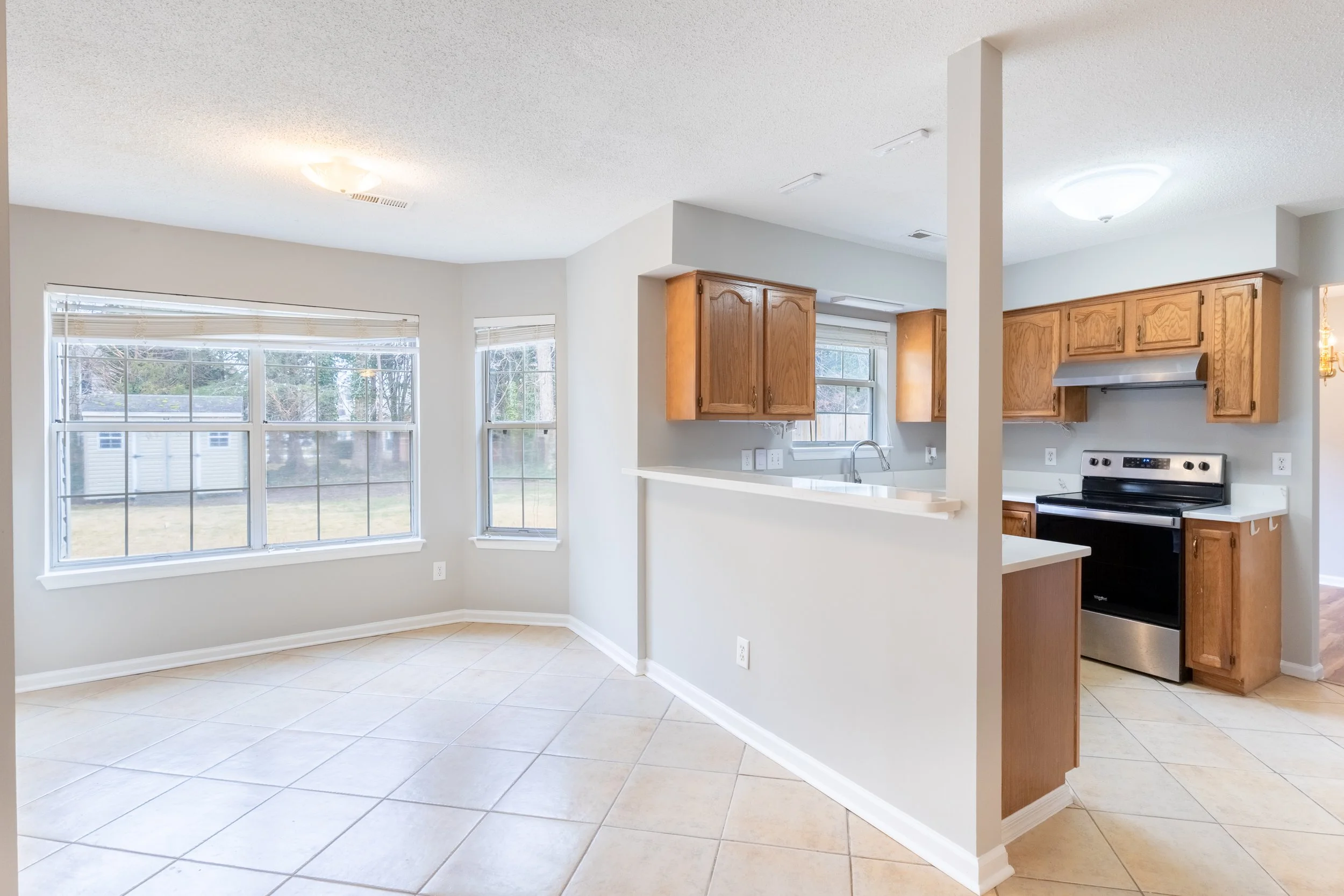 Empty kitchen and dining area with large windows, wooden cabinets, black stove, and tiled floor.