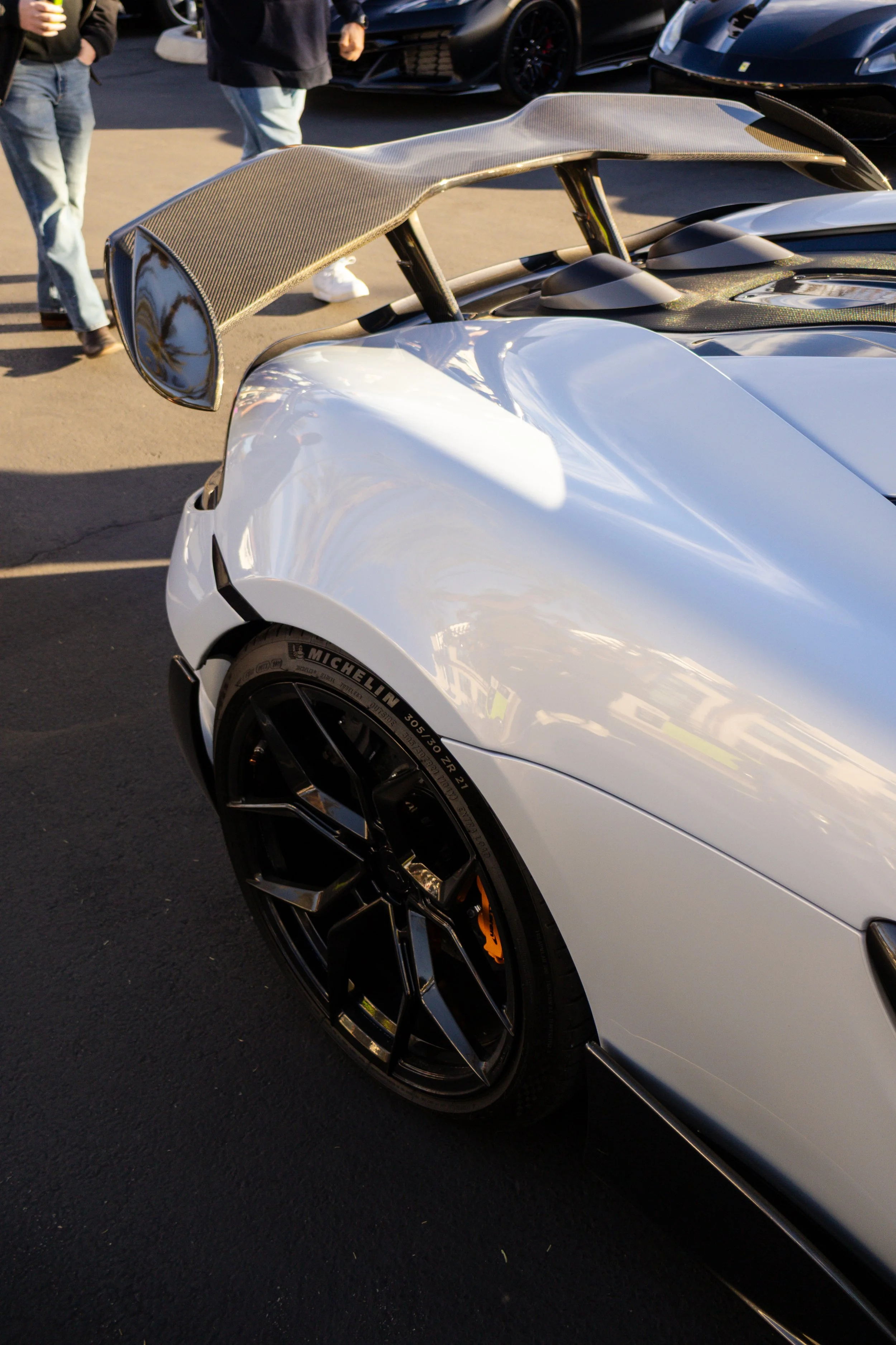 Close-up of the front corner of a white luxury sports car with a prominent carbon fiber rear wing and black wheels with Michelin tires, parked on a black asphalt surface with people in the background. Car meet. Cars and coffee. Rollers. Mclaren.