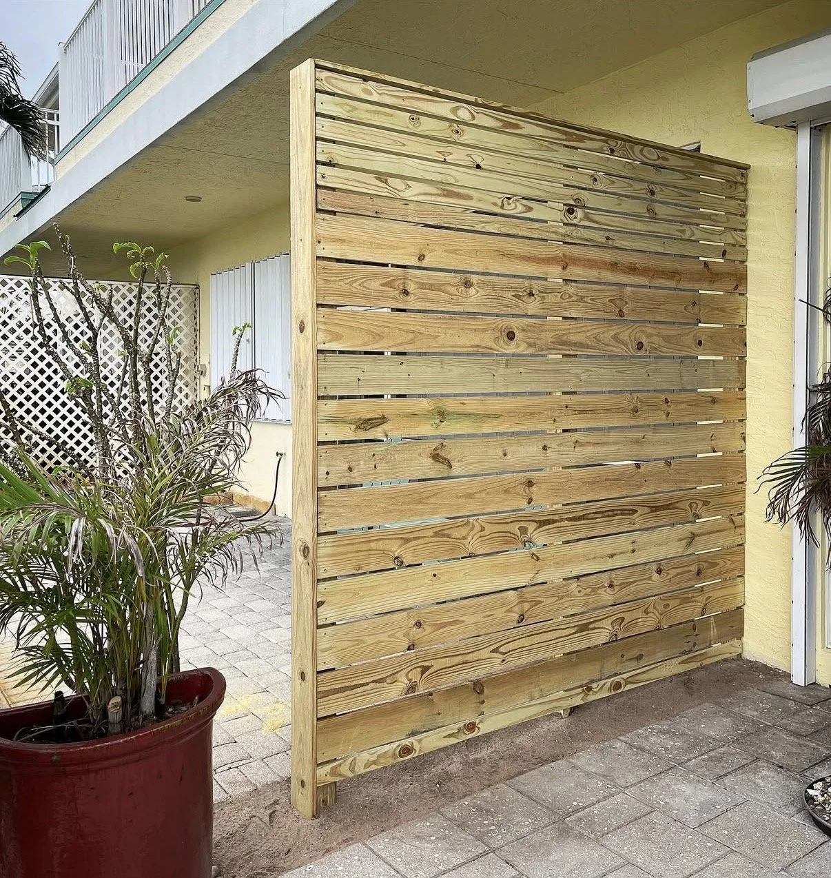 Image of a wooden privacy screen or fence outside a yellow house, next to potted plants on a patio.