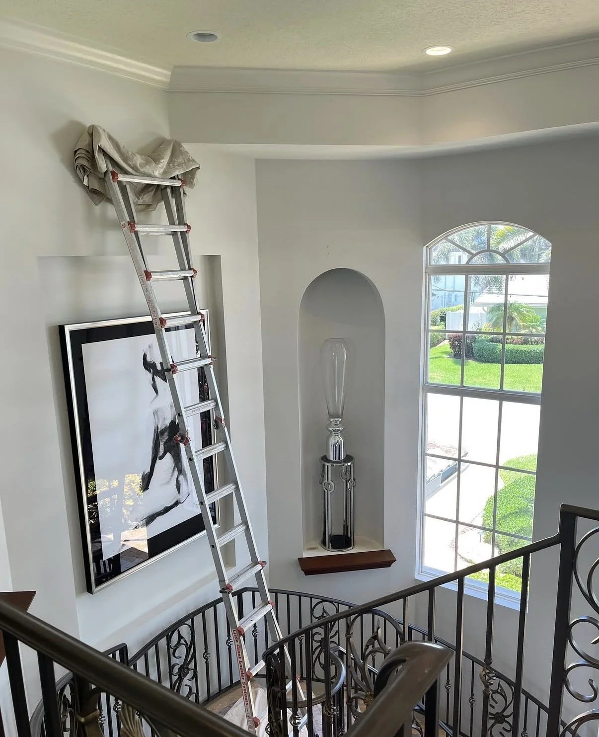 A ladder leaning against a white wall inside a house with a large window, a large framed art piece, and a decor tall glass sculpture on a stand.