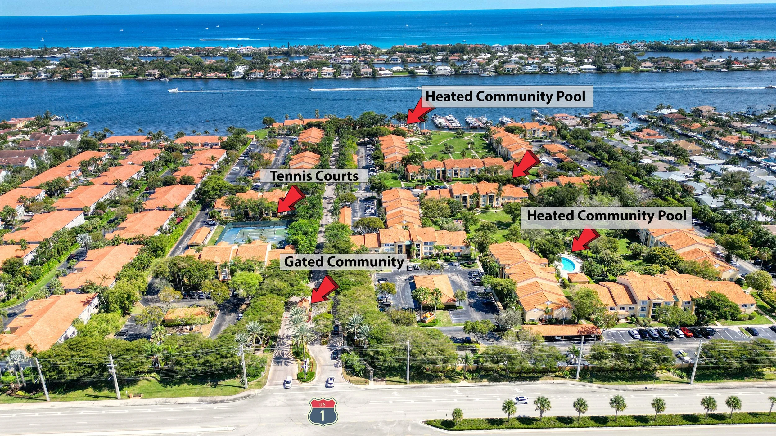 Aerial view of a residential community with multiple labeled amenities including Tennis Courts, Gated Community, Heated Community Pool, and a second Heated Community Pool, with water and boats in the background and palm trees along the street.