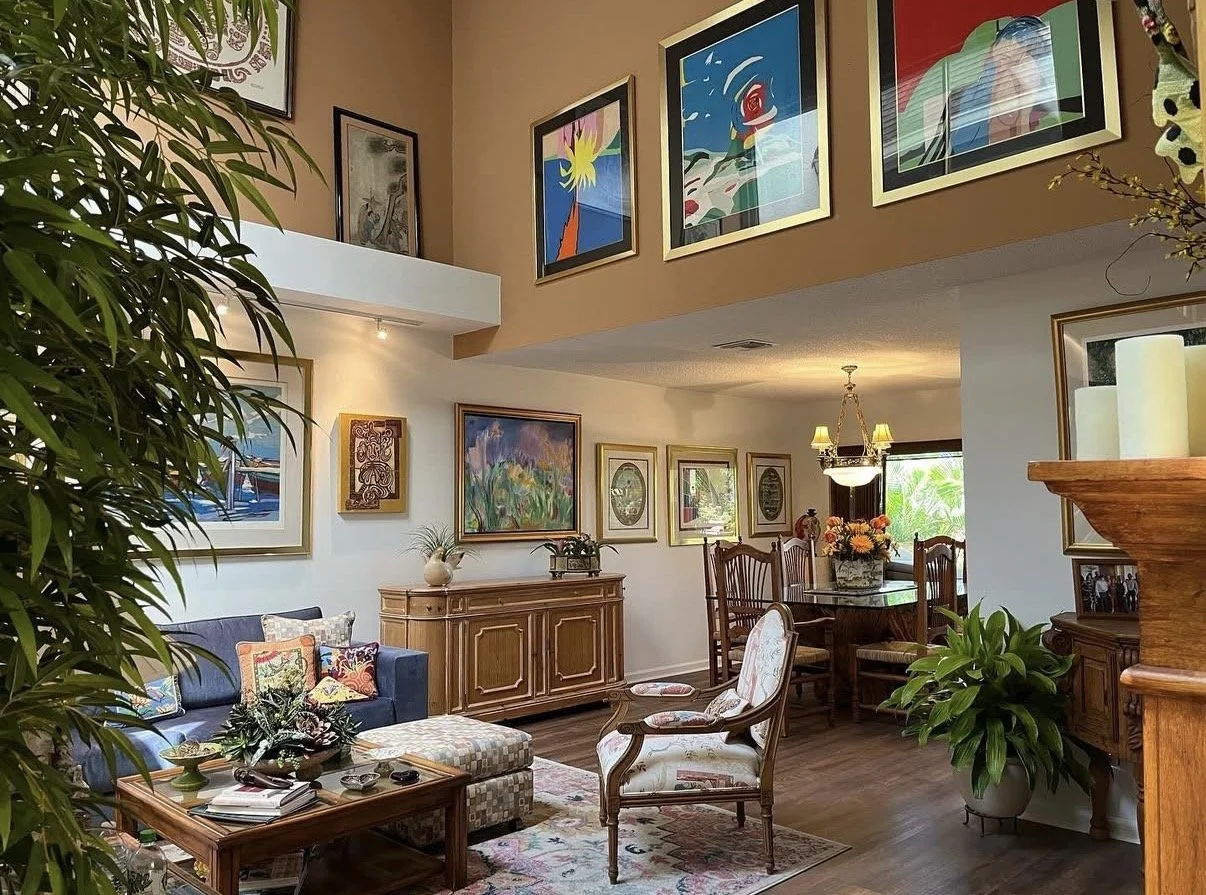 A living room with colorful artwork on the walls, a blue sofa with decorative pillows, a wooden cabinet, an armchair, a dining table with chairs, and several potted plants.