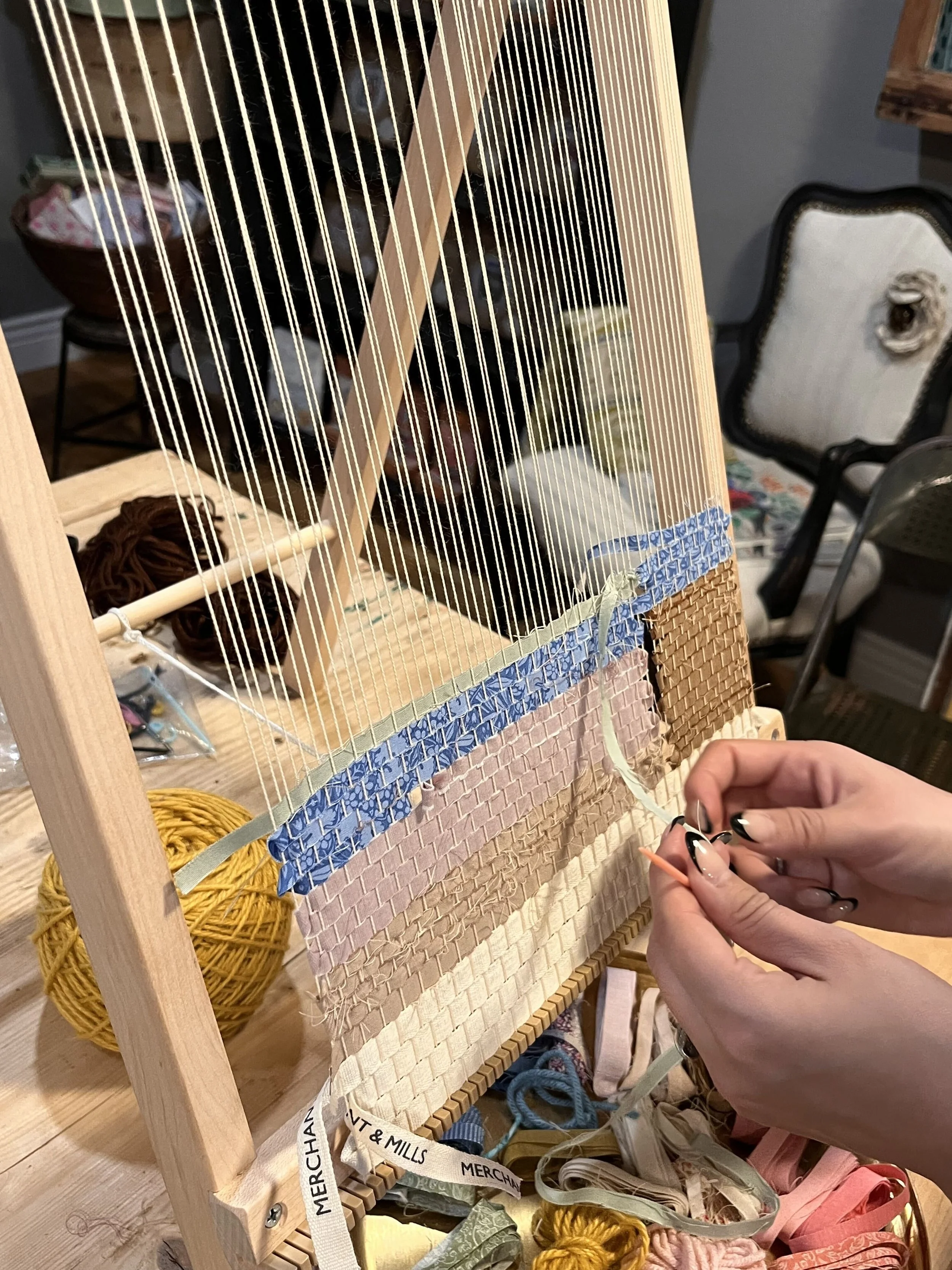 A crafter weaving on a community loom with ribbons in shades of blue, mauve, tan, off-white, and sage green.