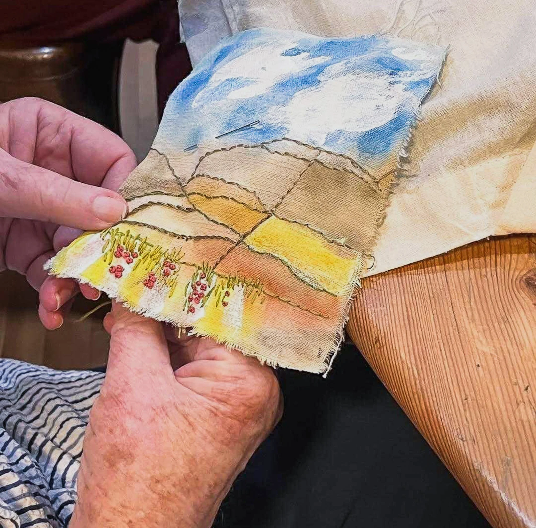 Someone embroidering a patch of textured fabric painted with a landscape using earth tones and blues