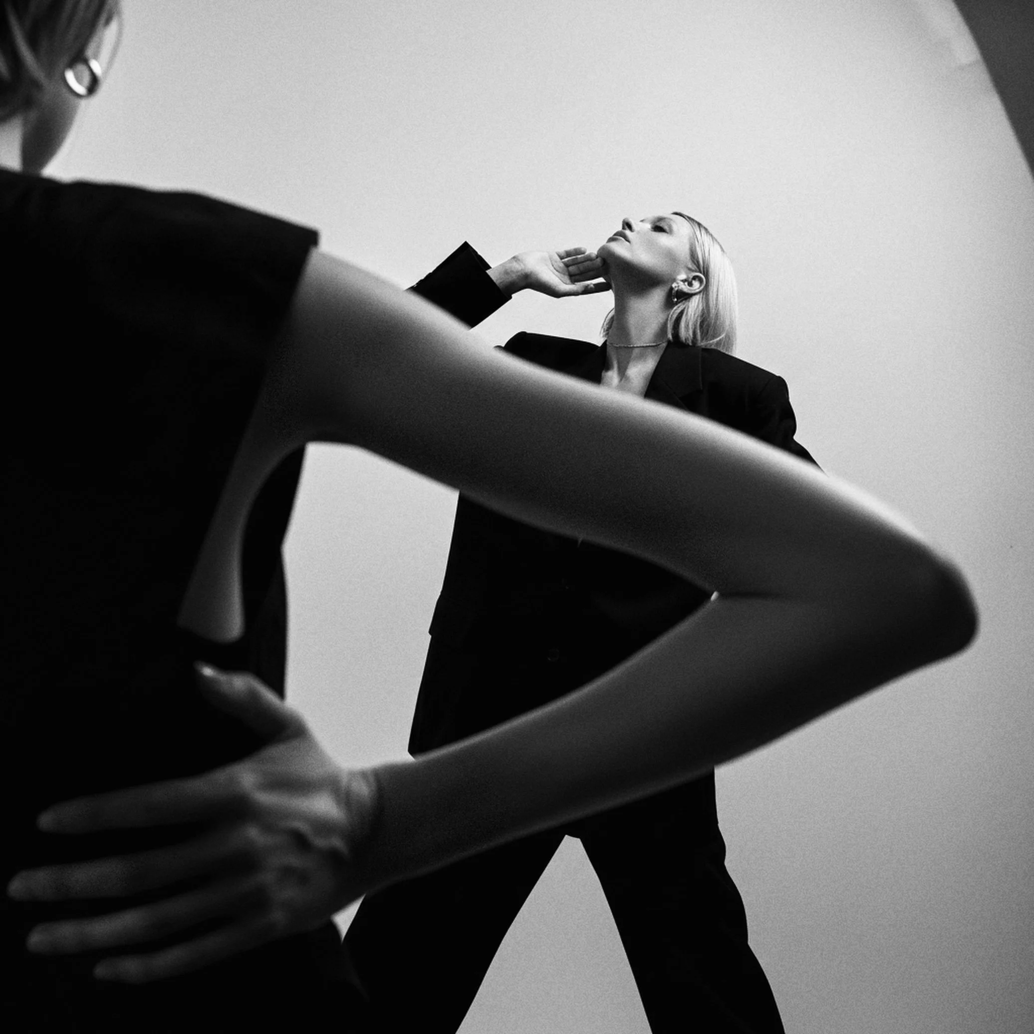 Black and white photo of a woman in professional attire posing with her hand on her neck, seen from a low angle, with another person's arm in the foreground.