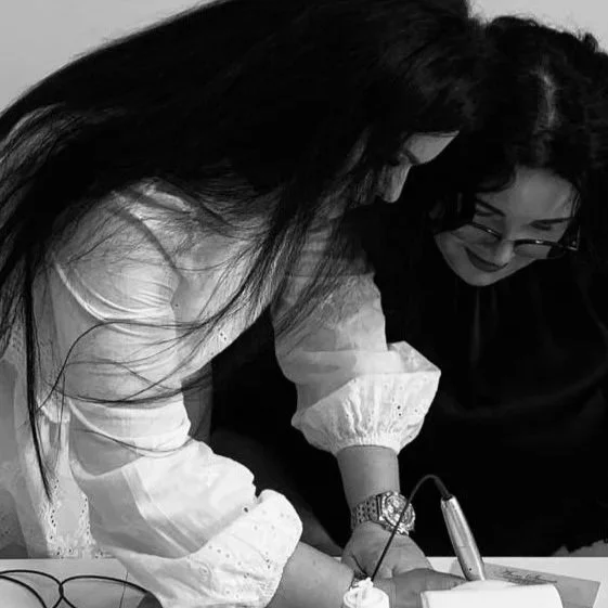 Two women with long hair and glasses, one in a white top and the other in a dark top, working together at a table, possibly on a project or craft.