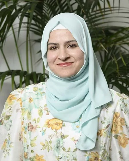 A woman wearing a light blue hijab and a floral blouse, standing in front of green plants.