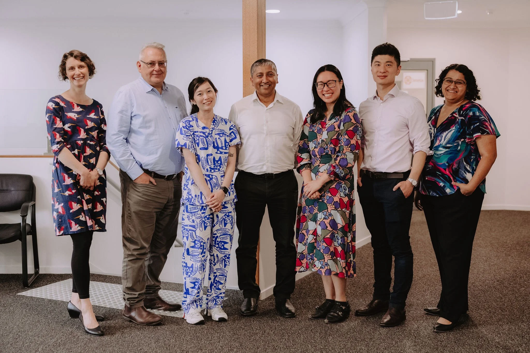 Seven diverse professionals standing in a row inside an office, smiling at the camera, dressed in casual business attire.