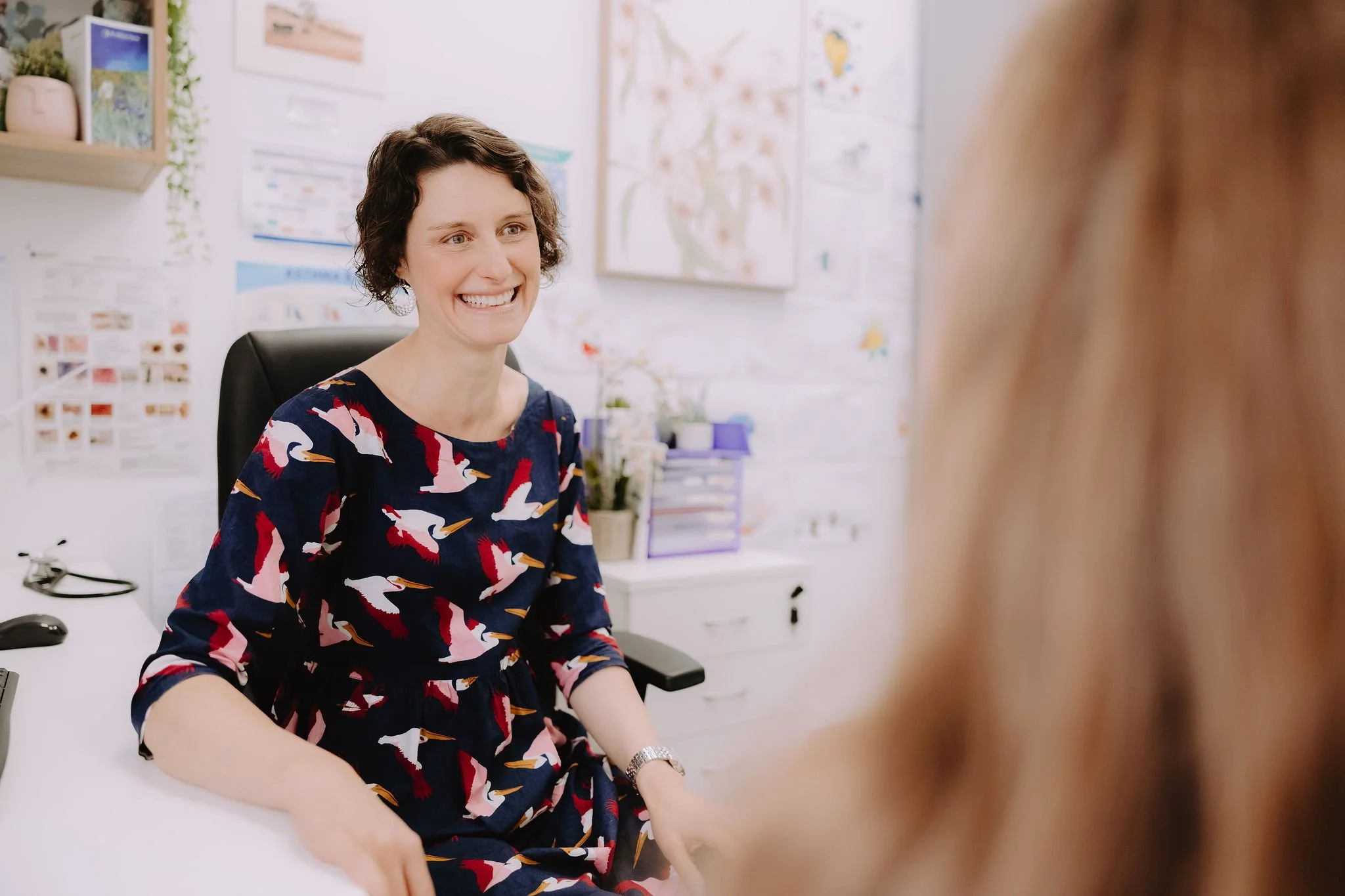 A woman with short curly dark hair and a striped navy dress with pink, white, and orange flamingos is sitting in a chair in a doctor's office, smiling while talking to a patient with blonde hair.