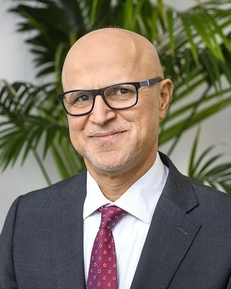 A bald man wearing glasses, a dark suit, white shirt, and maroon tie, standing in front of green plants.