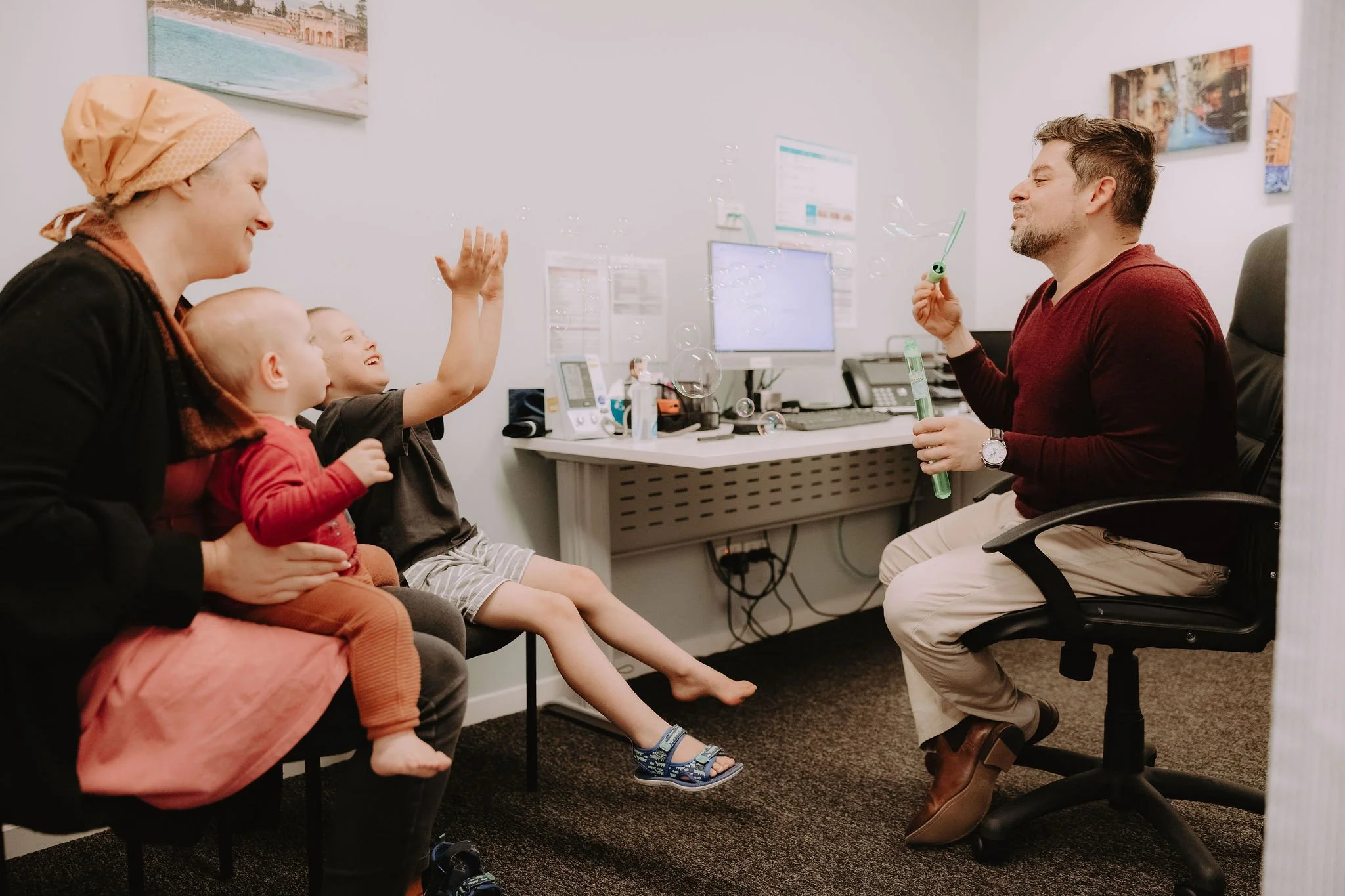 A woman with a headscarf and two children sitting in an office, children playing with bubbles blown by a man sitting in front of them at a desk. The children are smiling and reaching for the bubbles.