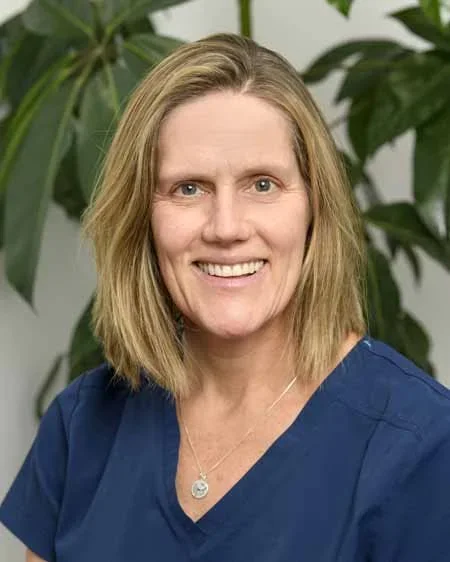 A smiling woman with shoulder-length blond hair wearing a blue top, standing in front of green leafy plants.