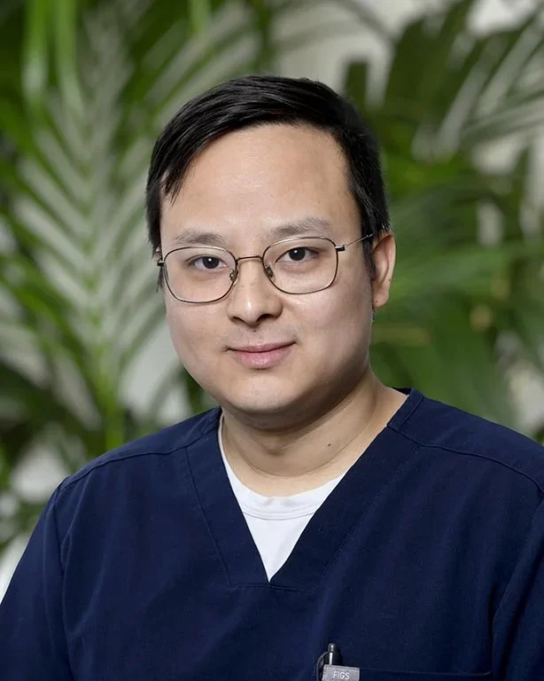 A man with glasses wearing navy blue medical scrubs in front of green plants.
