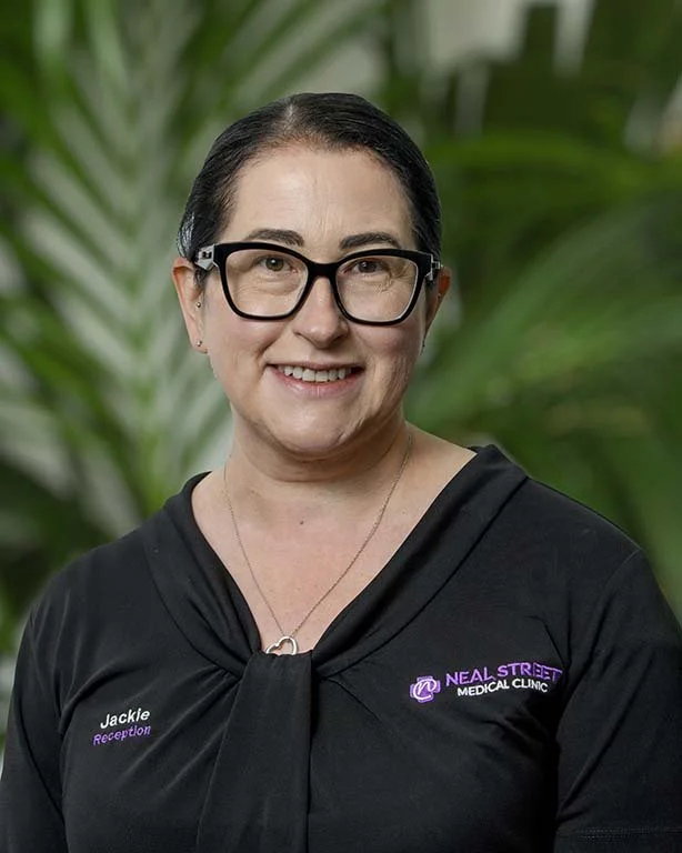 A woman with dark hair, glasses, and a black top with a logo that says 'Neal Street Medical Clinic,' standing in front of green plants.