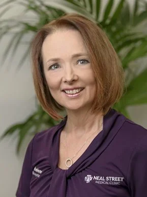 A woman with shoulder-length red hair smiling, wearing a purple Neal Street Medical Clinic scrub top, standing in front of green plants.