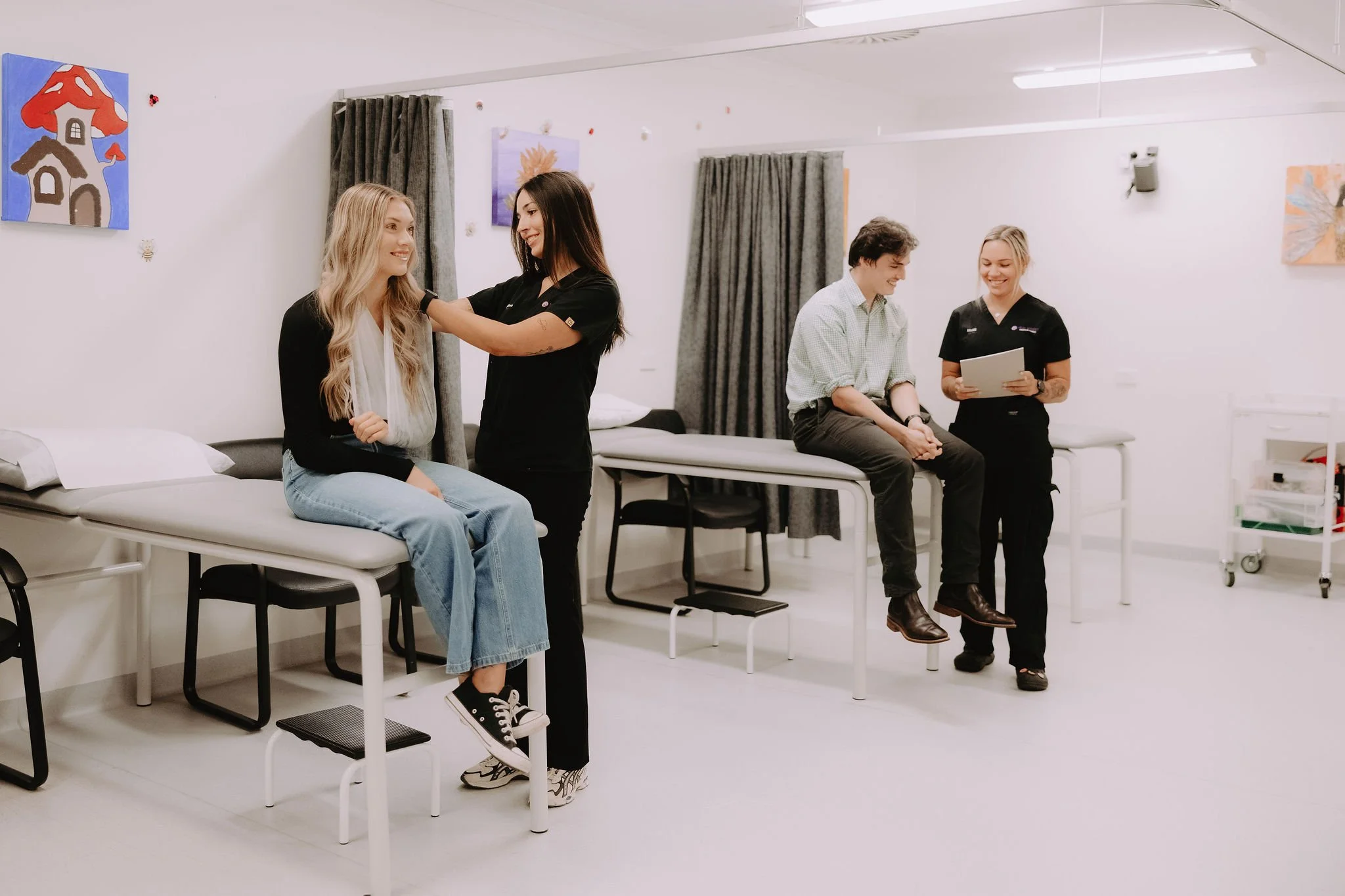 Two women are in a medical examination room, with one sitting on an examination table and the other standing beside her. In the background, a man and a woman are seated on examining tables, with the woman holding a clipboard. The room has white walls, curtains, and colorful wall art.