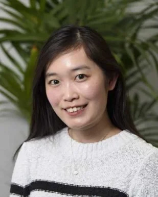 A young woman with long dark hair smiling in front of large green plants.
