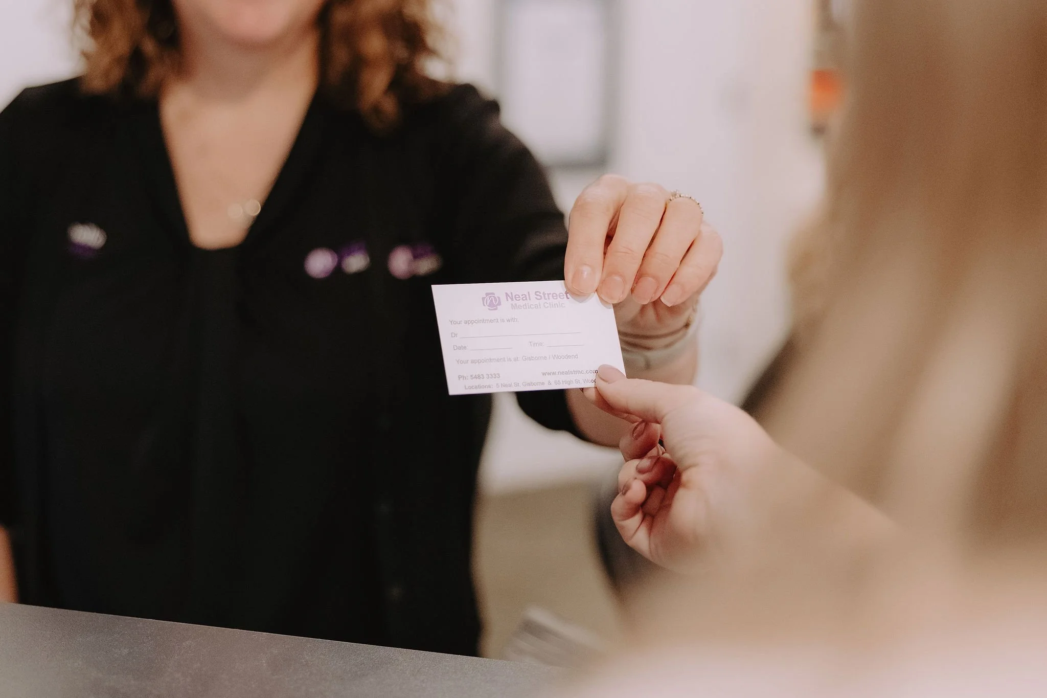 A woman handing a small medical appointment card to another person in a reception area.