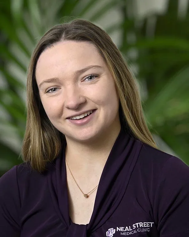 Professional woman with shoulder-length light brown hair, smiling, wearing a dark purple top with a necklace, standing against a green leafy background.