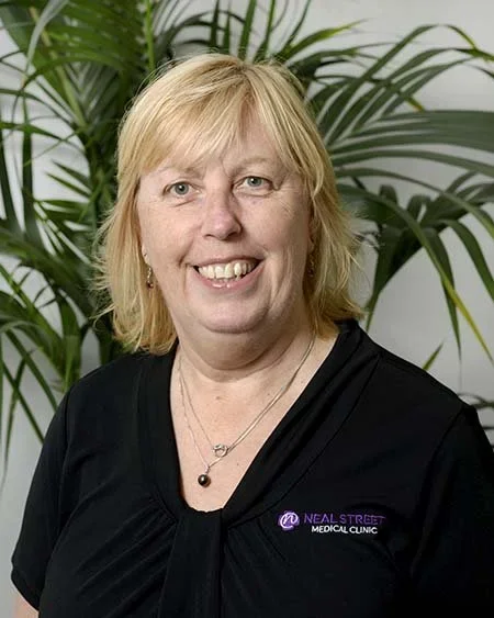 A smiling woman with blonde hair wearing a black shirt with 'Neal Street Medical Clinic' logo, standing in front of green plants.