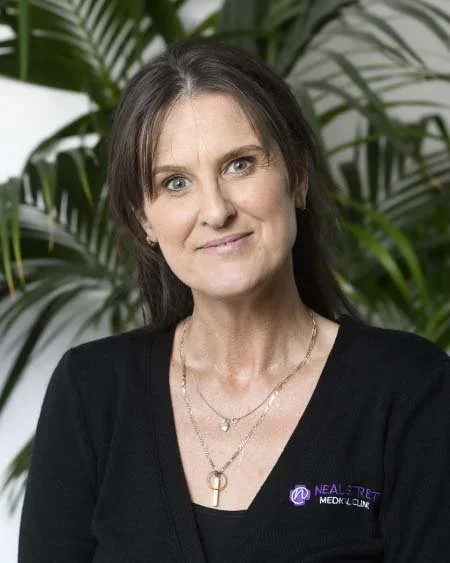 A woman with shoulder-length dark hair wearing a black top with a purple logo, silver necklace, in front of green tropical plants.