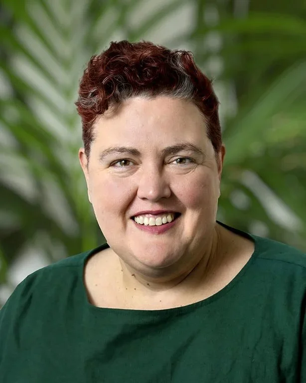 Portrait of a smiling woman with short, curly red hair, wearing a green top, with a blurred green foliage background.