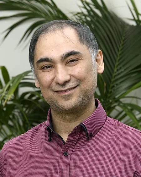 A man with short dark hair, wearing a maroon button-up shirt, smiling while standing in front of green palm leaves.