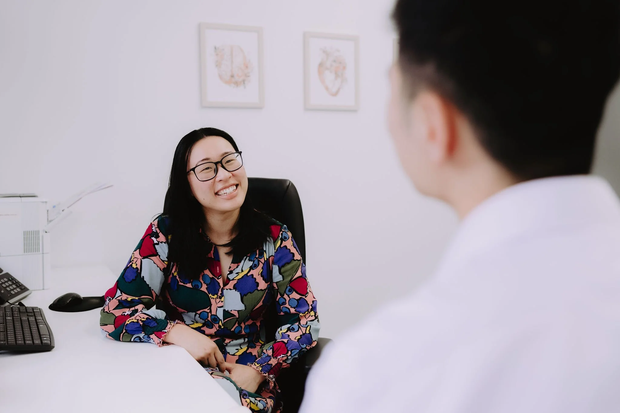 A woman with glasses and a colorful floral blouse smiling and talking to a man in a white shirt in an office setting.