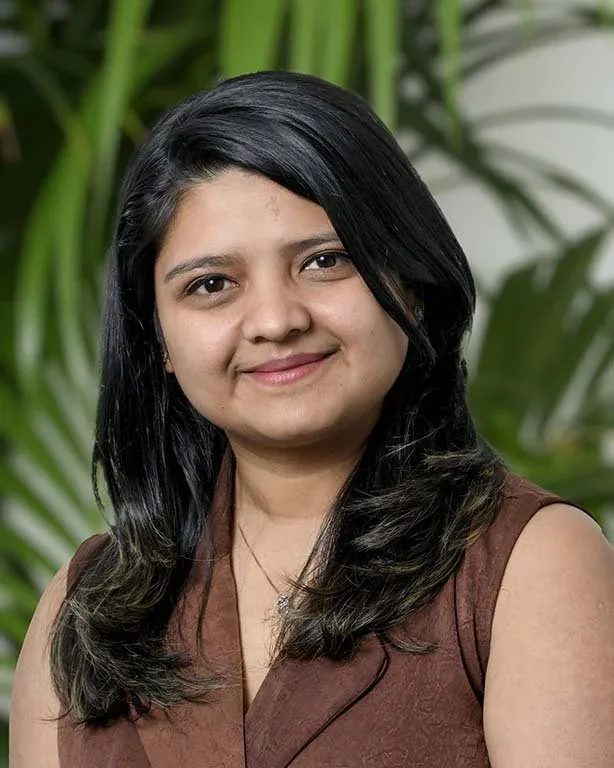 A woman with black hair and a slight smile posing in front of a green leafy background.