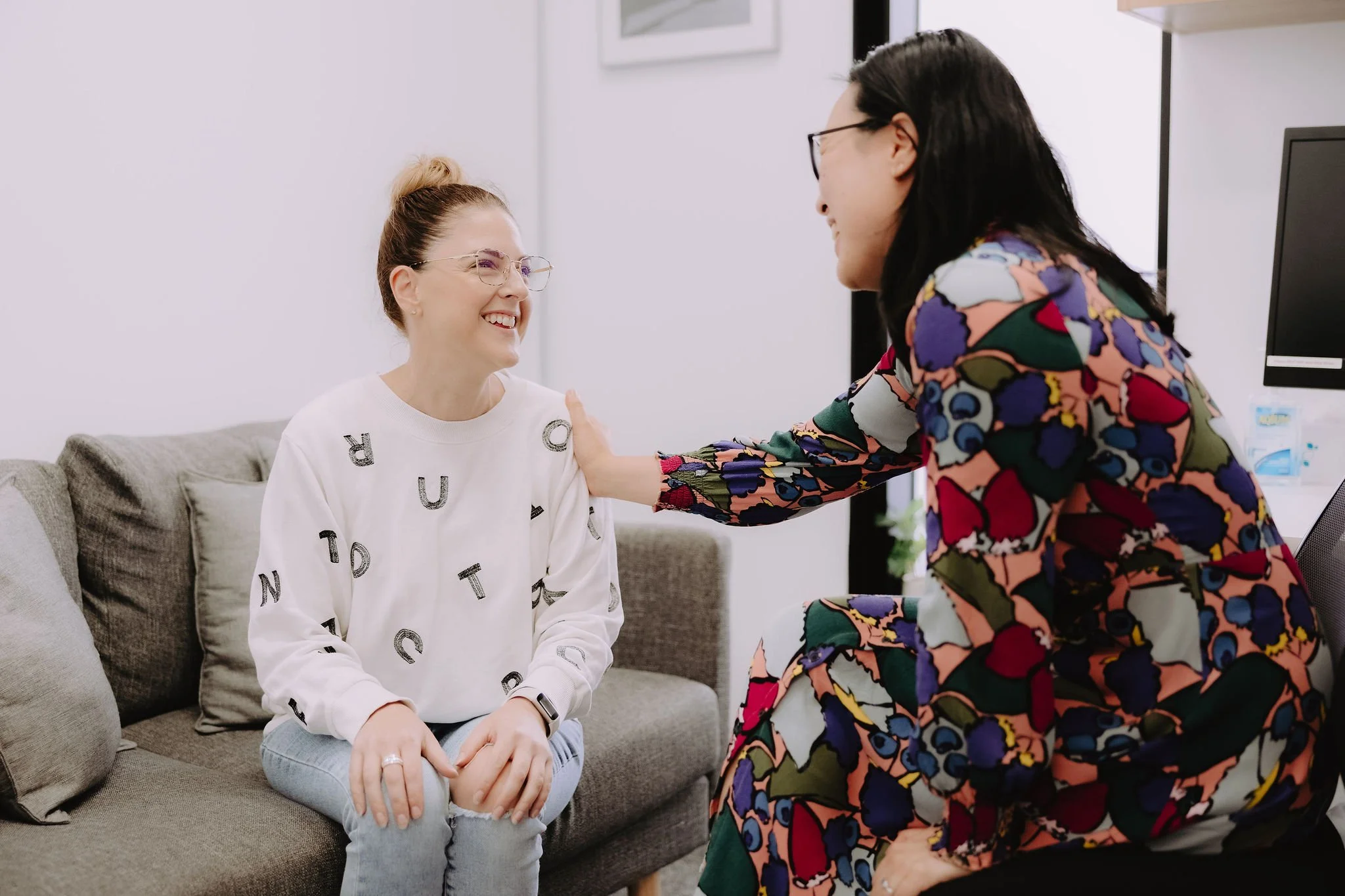 A woman with glasses and a white sweater with alphabet letters sits on a gray couch, smiling at another woman with glasses and a colorful floral dress. The woman in floral dresses her hand on the other woman's shoulder, and they seem to be having a friendly, positive interaction in a well-lit room.