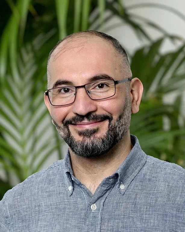 A man with glasses and a beard smiling, wearing a blue button-up shirt, with green plants in the background.